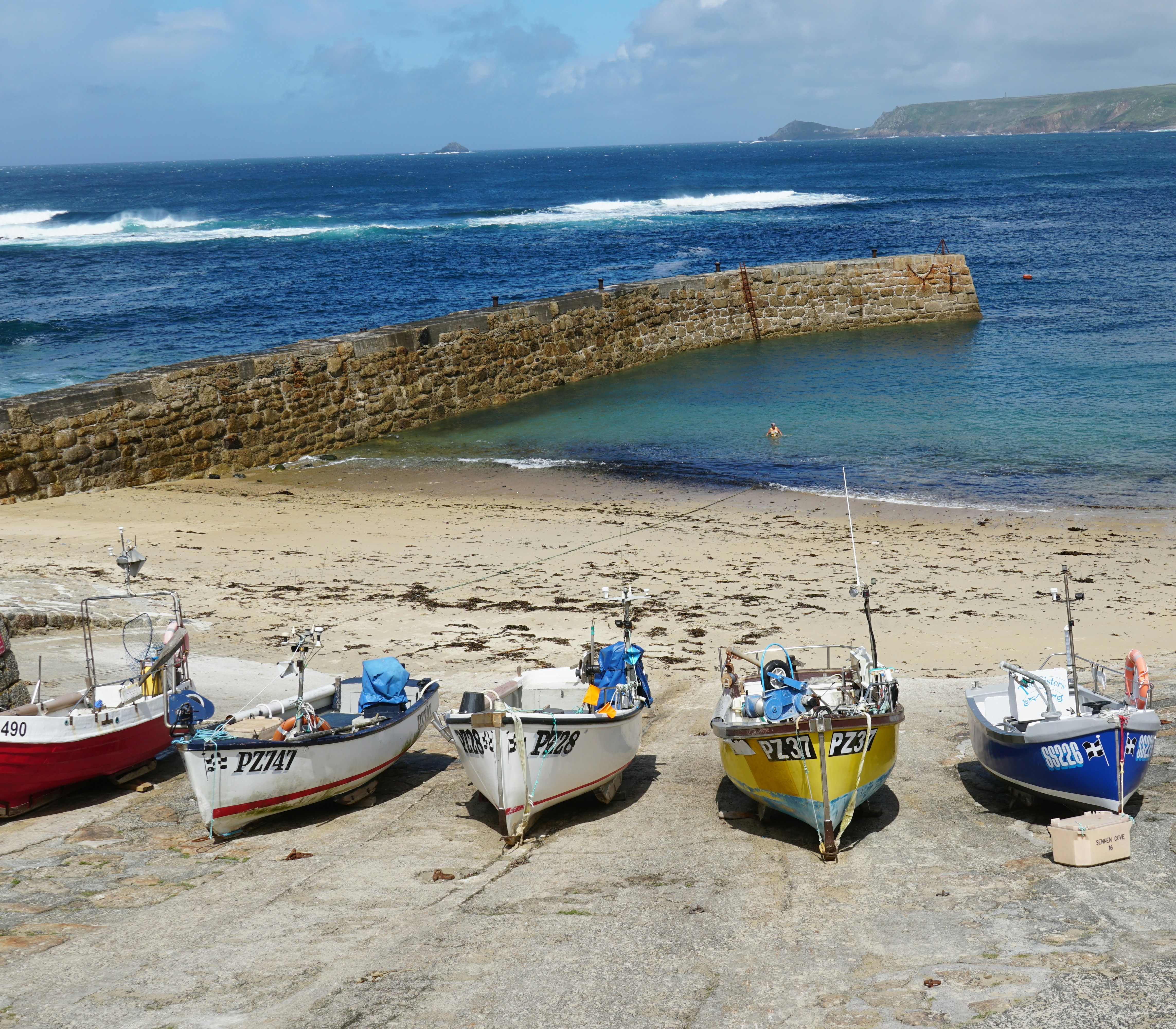 Small fishing boats lined up on a slipway near a sandy beach and a stone harbour wall with the sea in the background.