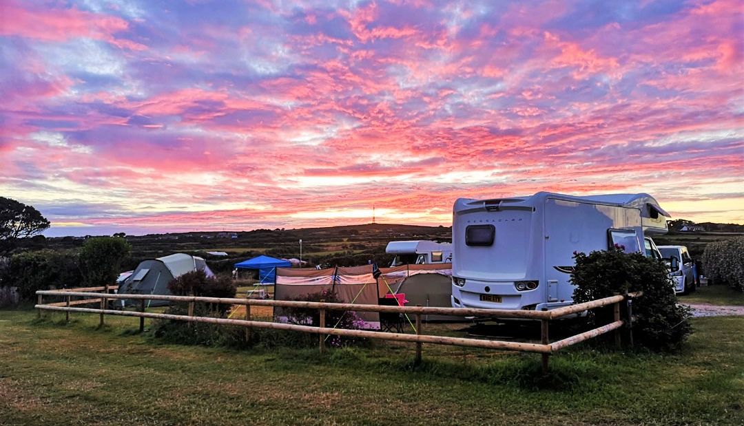 A vibrant sunrise over a campsite with tents, a camper van, and grassy fields.