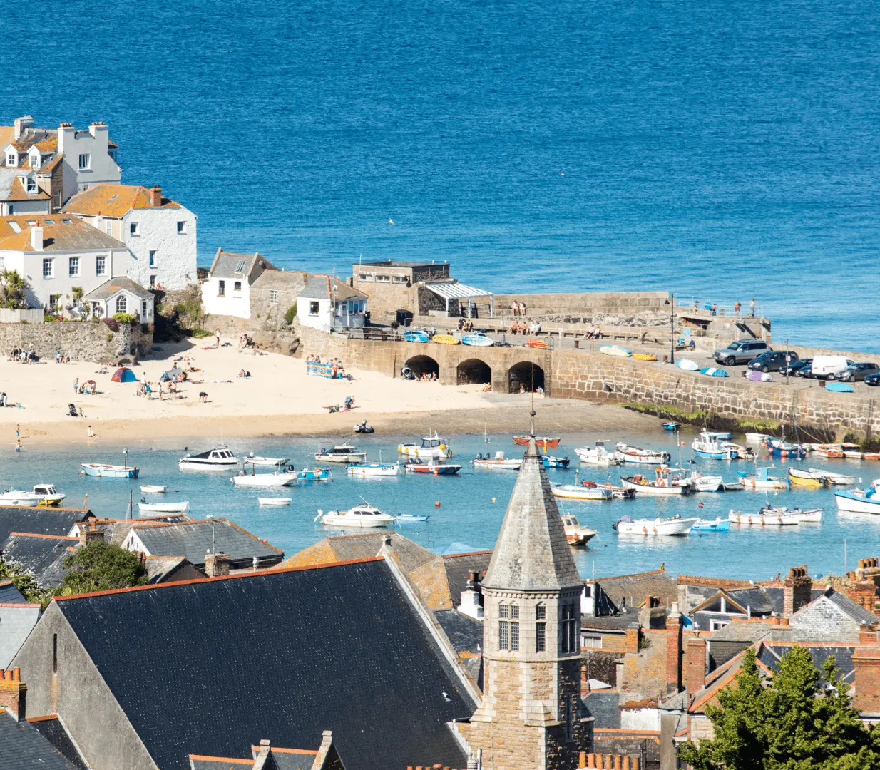 A picturesque coastal town with sandy beach, numerous small boats in the harbor, and quaint houses with orange-tiled roofs.