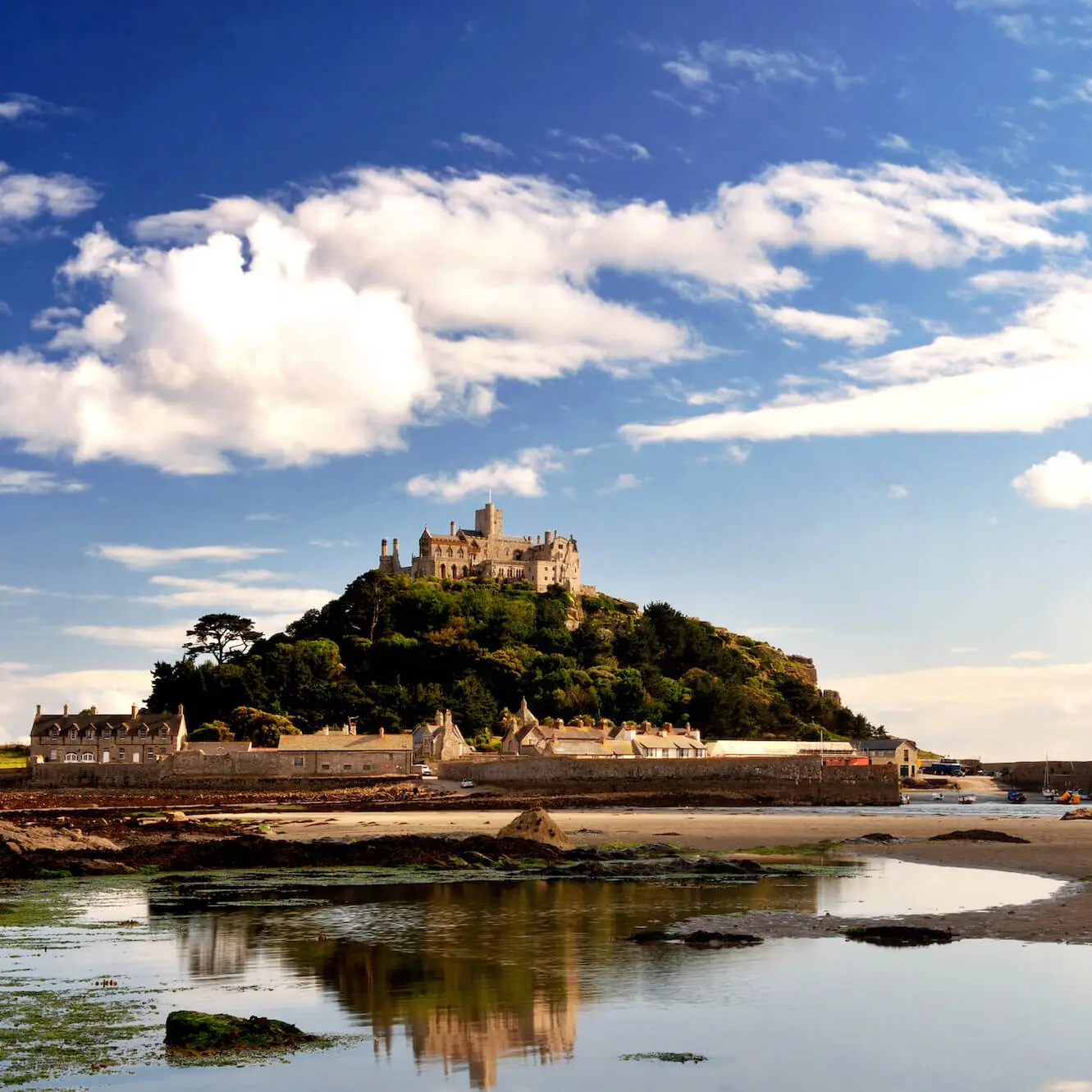 A castle on a hill with a village below, surrounded by water and a sandy shore under a blue sky with clouds.