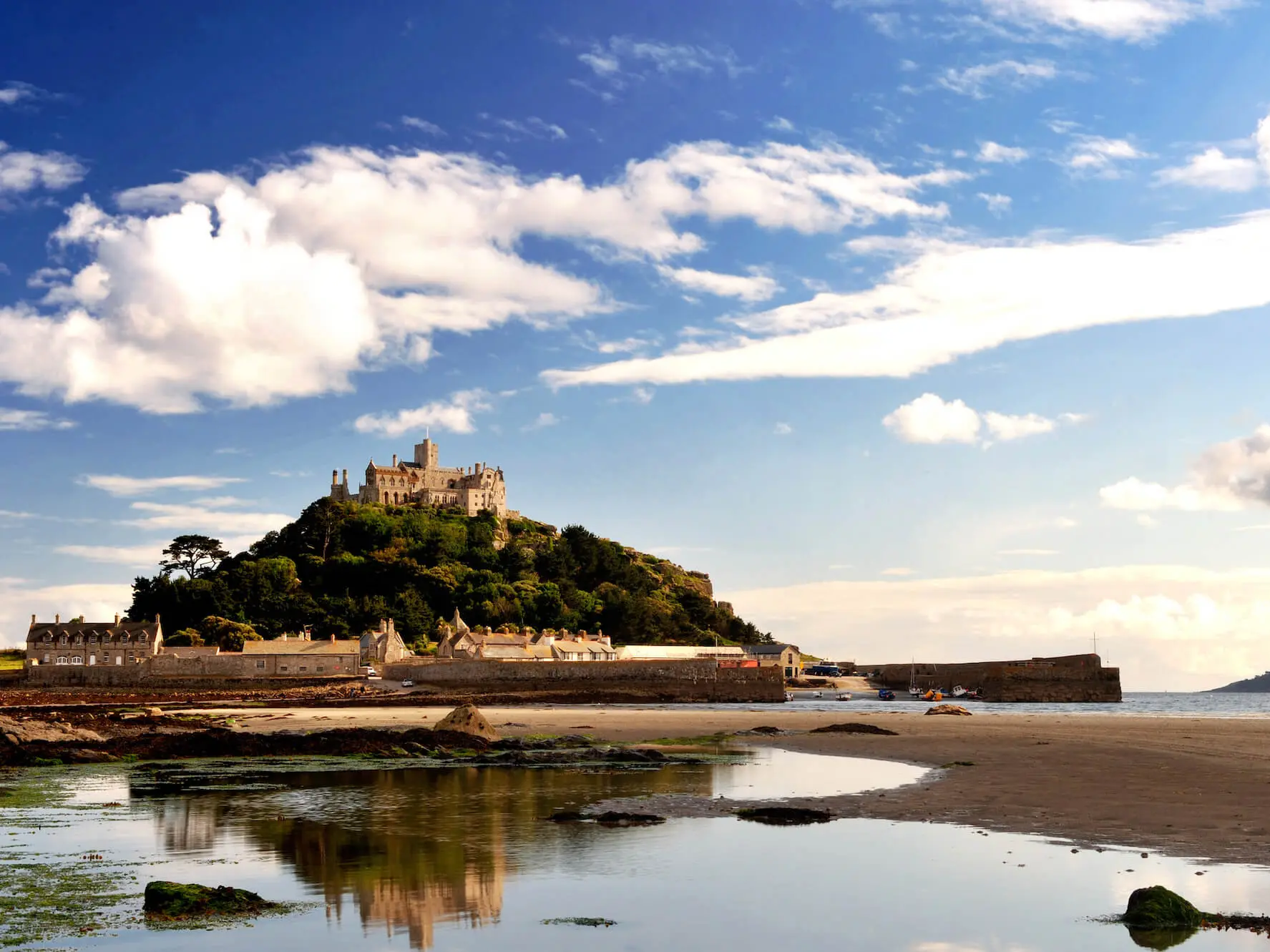 A castle on a hill with a village below, surrounded by water and a sandy shore under a blue sky with clouds.