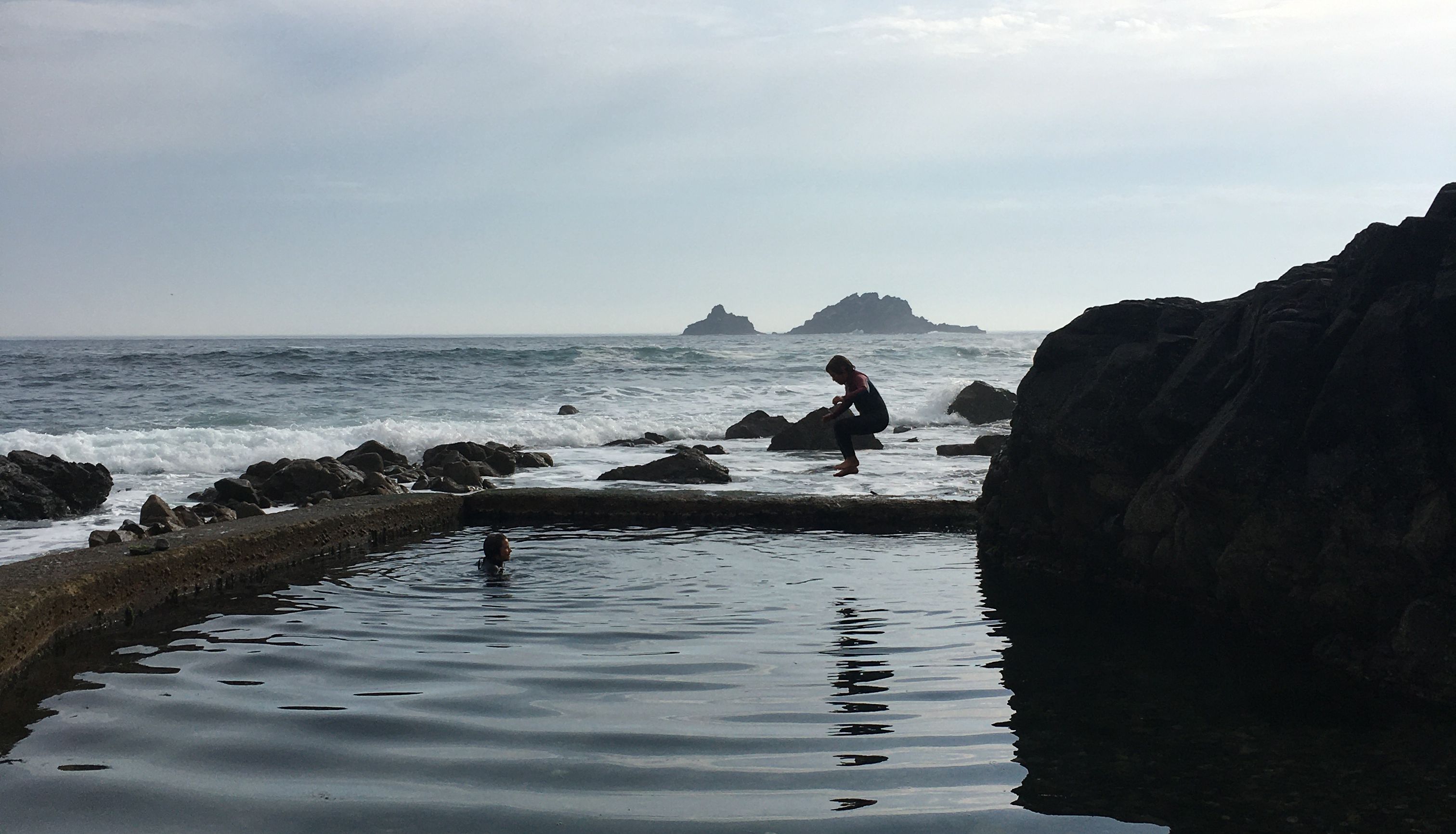 Person sitting on rocks beside a tidal pool with another person swimming, ocean and rocky islands in the distance