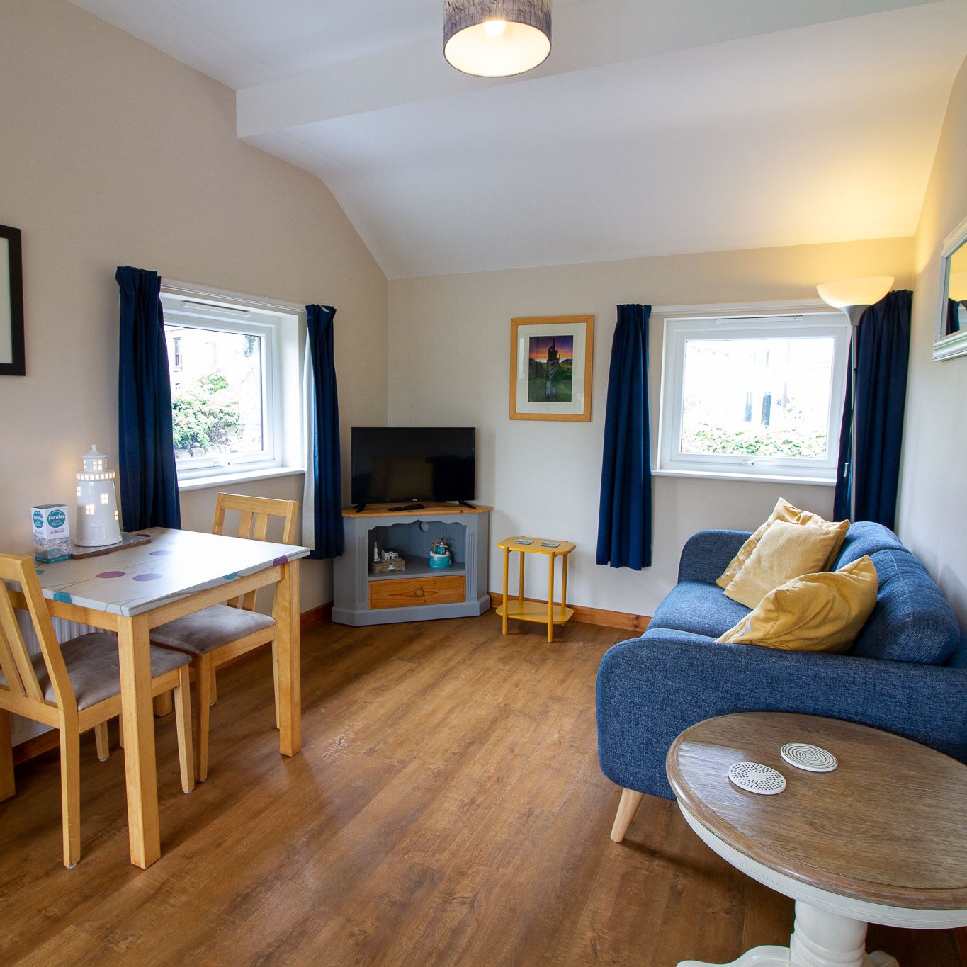 Cozy living and dining area with a blue sofa, wooden table and chairs, flat screen TV, and windows with blue curtains.