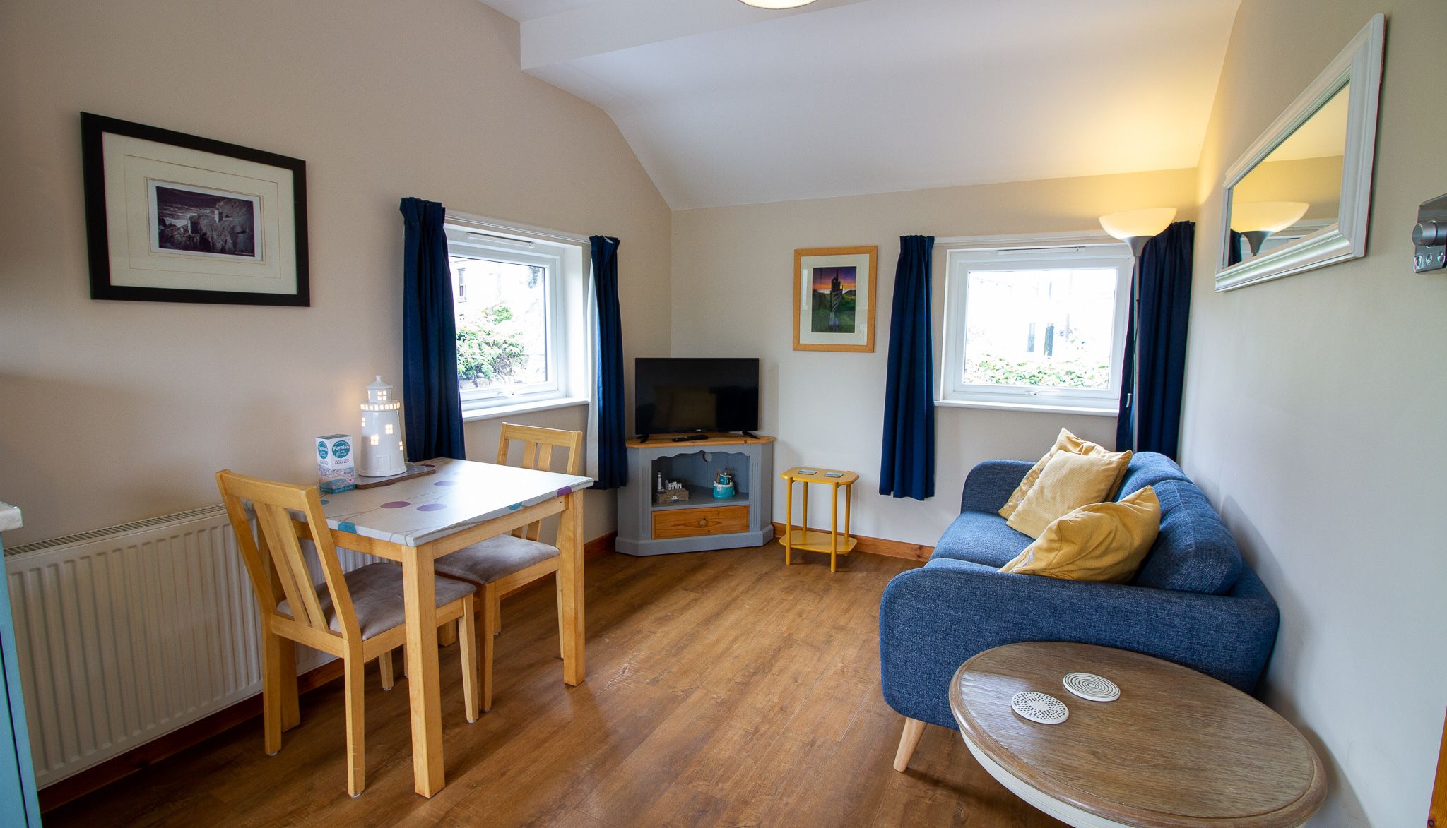 Cozy living and dining area with a blue sofa, wooden table and chairs, flat screen TV, and windows with blue curtains.