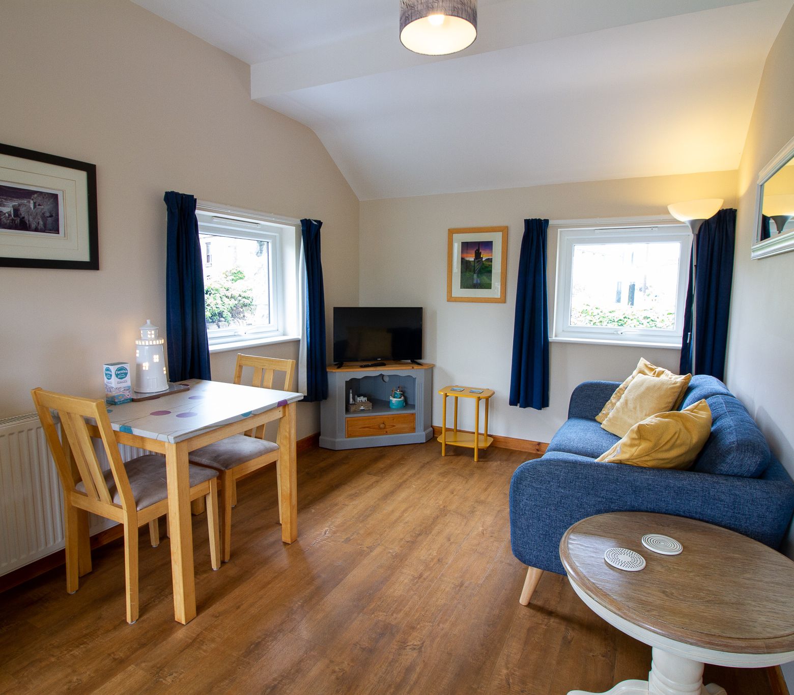 Cozy living and dining area with a blue sofa, wooden table and chairs, flat screen TV, and windows with blue curtains.