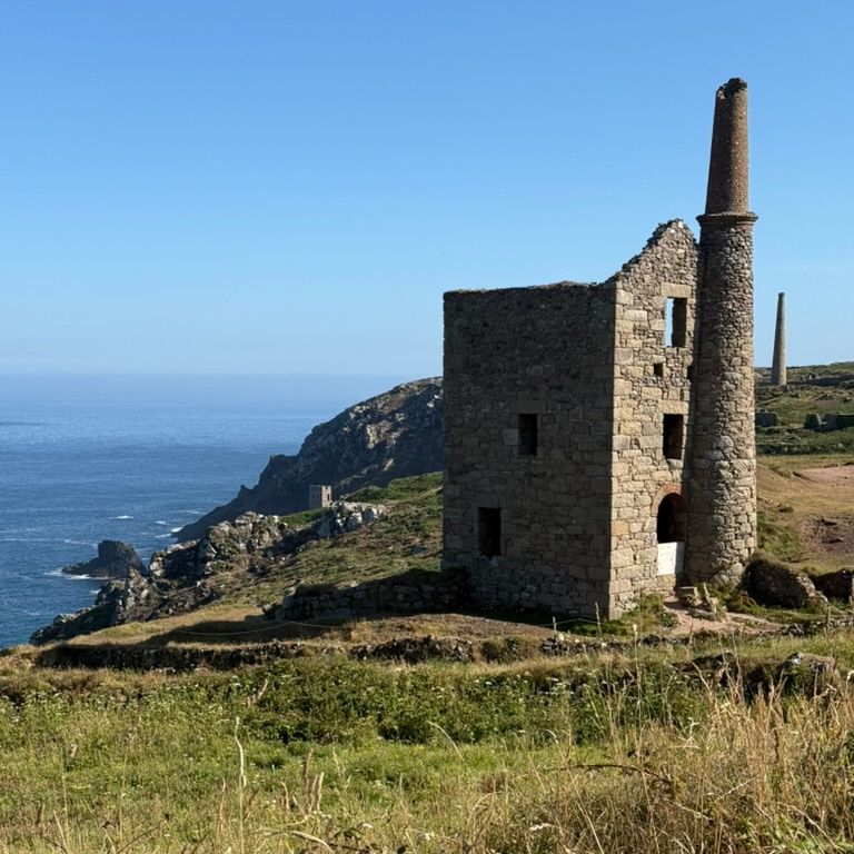 Old stone mining building with a tall chimney on a grassy cliff overlooking the sea.