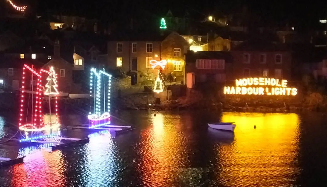 Colorful Christmas lights and illuminated sign reflecting on the water in Mousehole harbour at night.