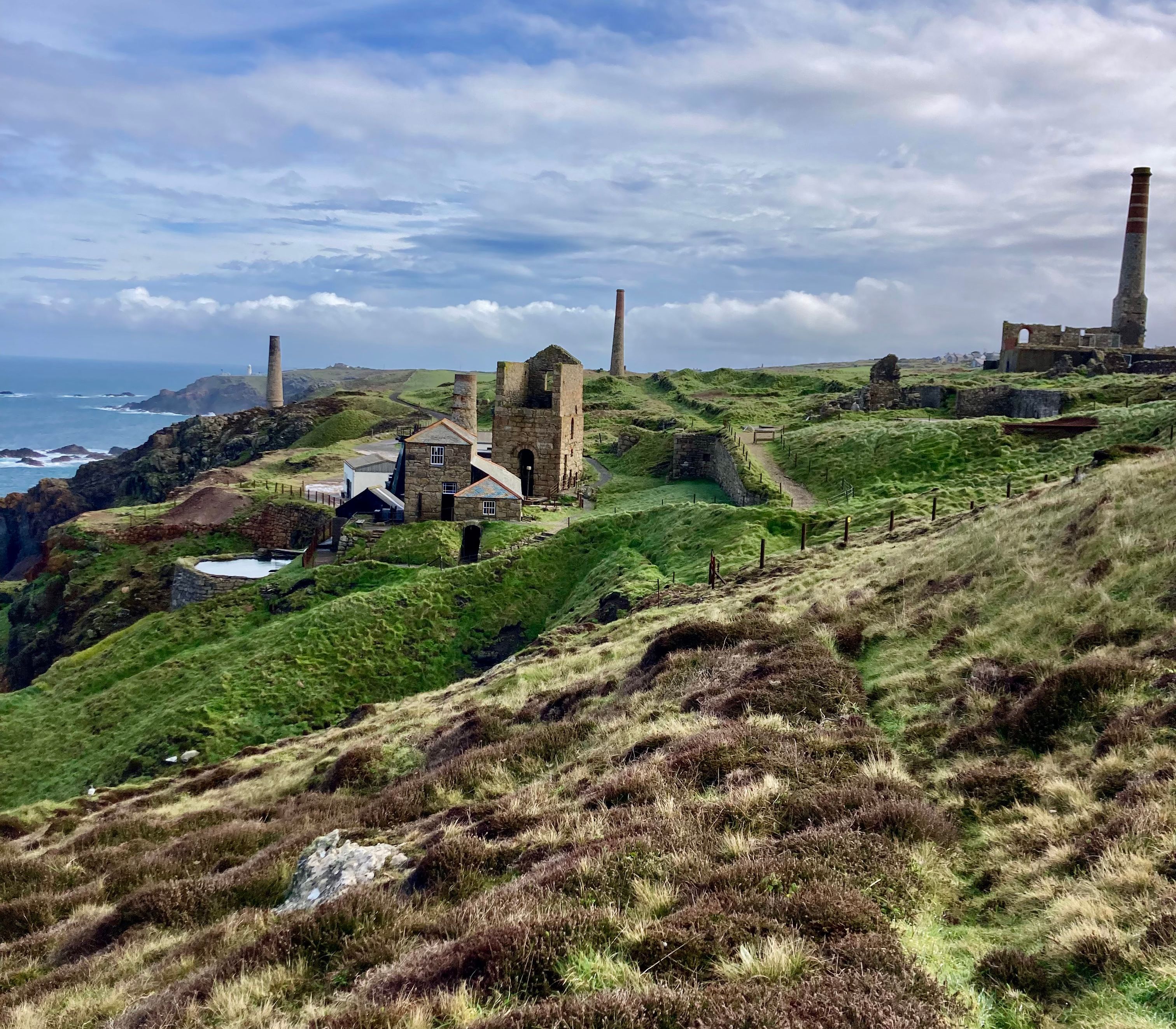 Ruins of old mining buildings with tall chimneys on a grassy coastal hill overlooking the sea under a cloudy sky.