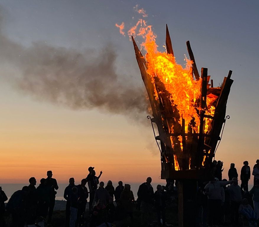 Large ceremonial beacon fire burning with people gathered around at sunset.
