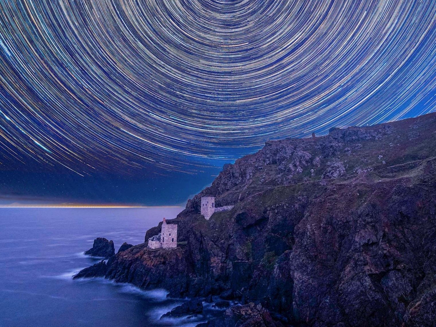 Long-exposure photo of star trails swirling above two stone towers on a rugged coastal cliff.