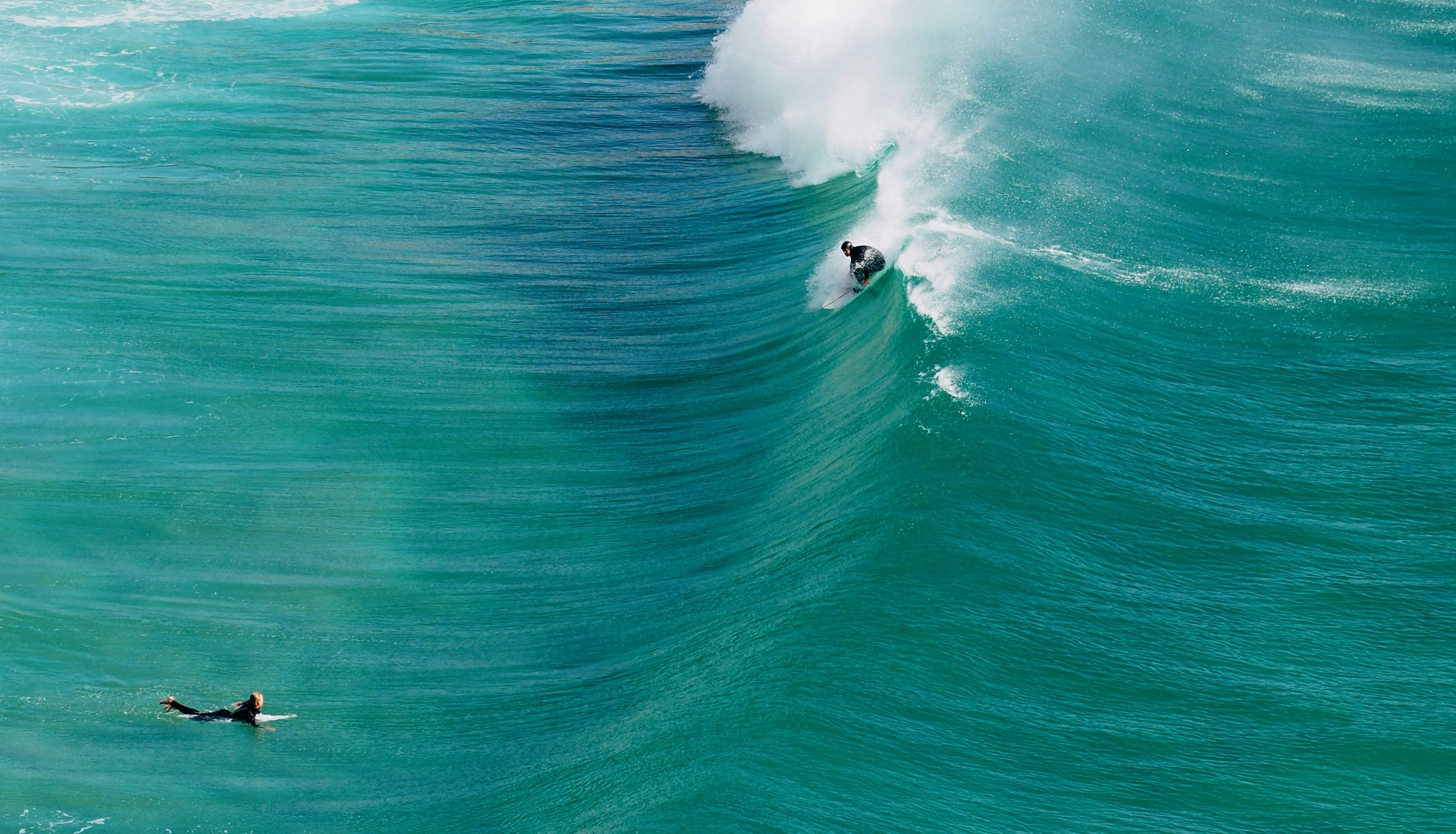Surfer riding a large turquoise wave with another surfer paddling nearby