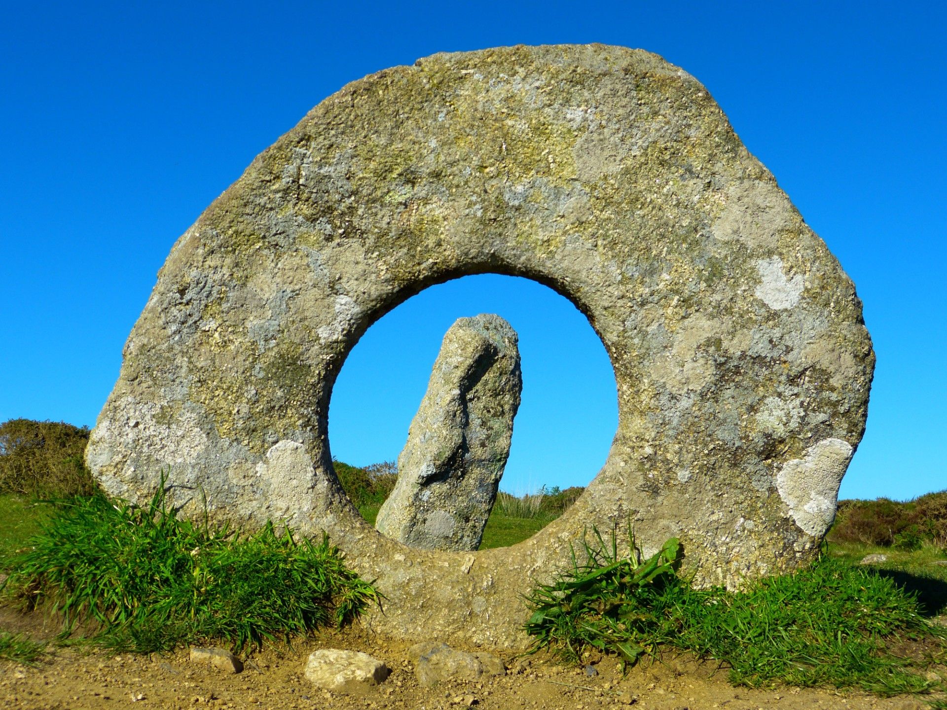 Large standing stone with a circular hole in the center and another upright stone visible through the hole, set against a bright blue sky.