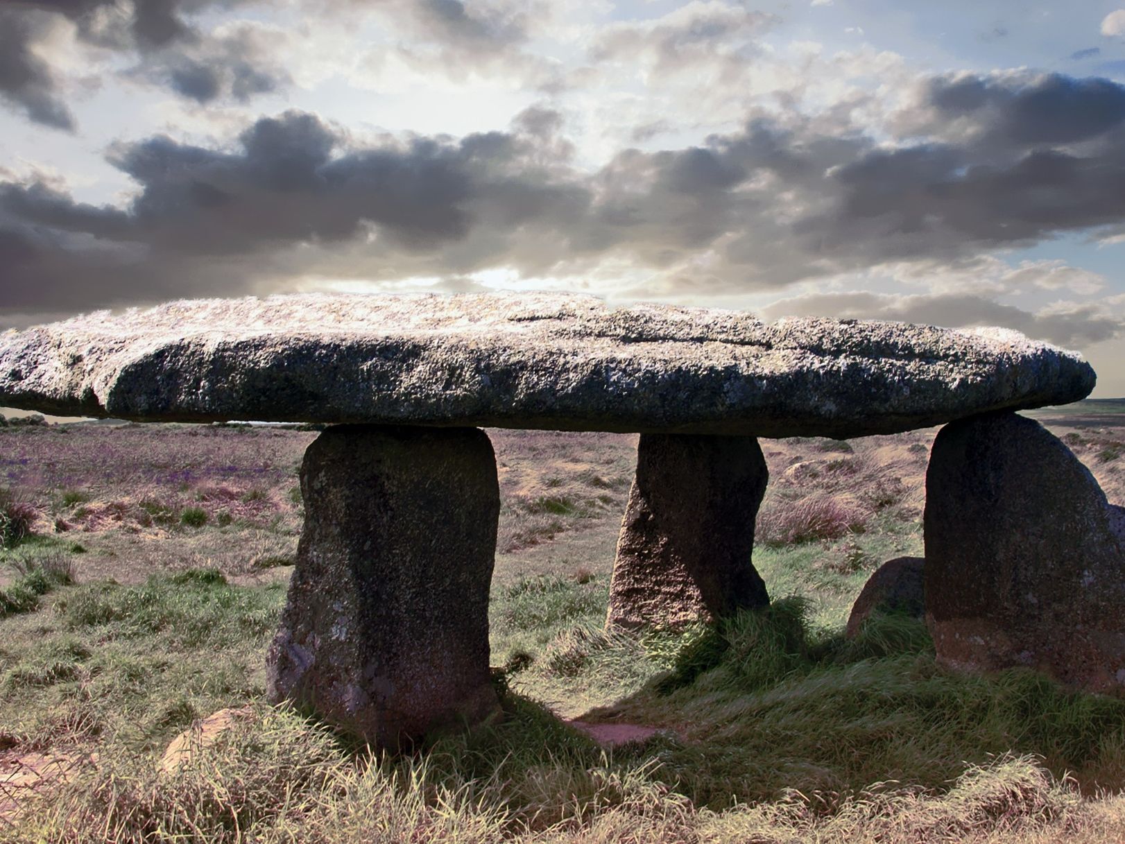Ancient stone dolmen structure with a large flat stone slab supported by upright stones in a grassy landscape under a cloudy sky.