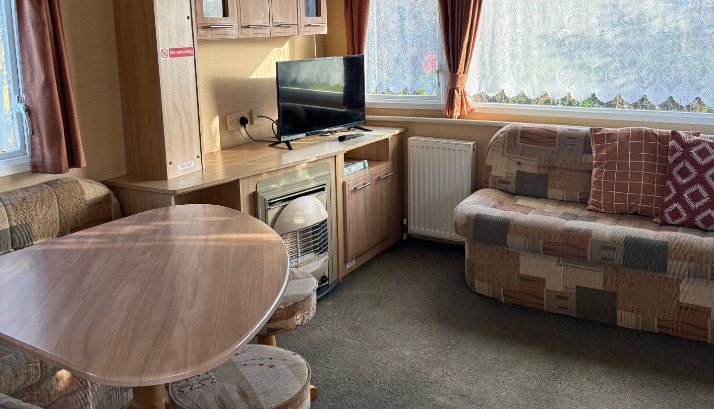 Cozy caravan living area with patterned sofas, a wooden dining table, TV, and large window with sheer curtains.