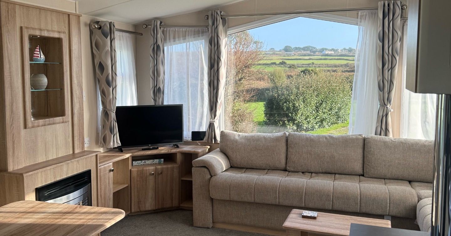 Modern caravan living room with beige sofa, TV, coffee table, and countryside view through large windows.