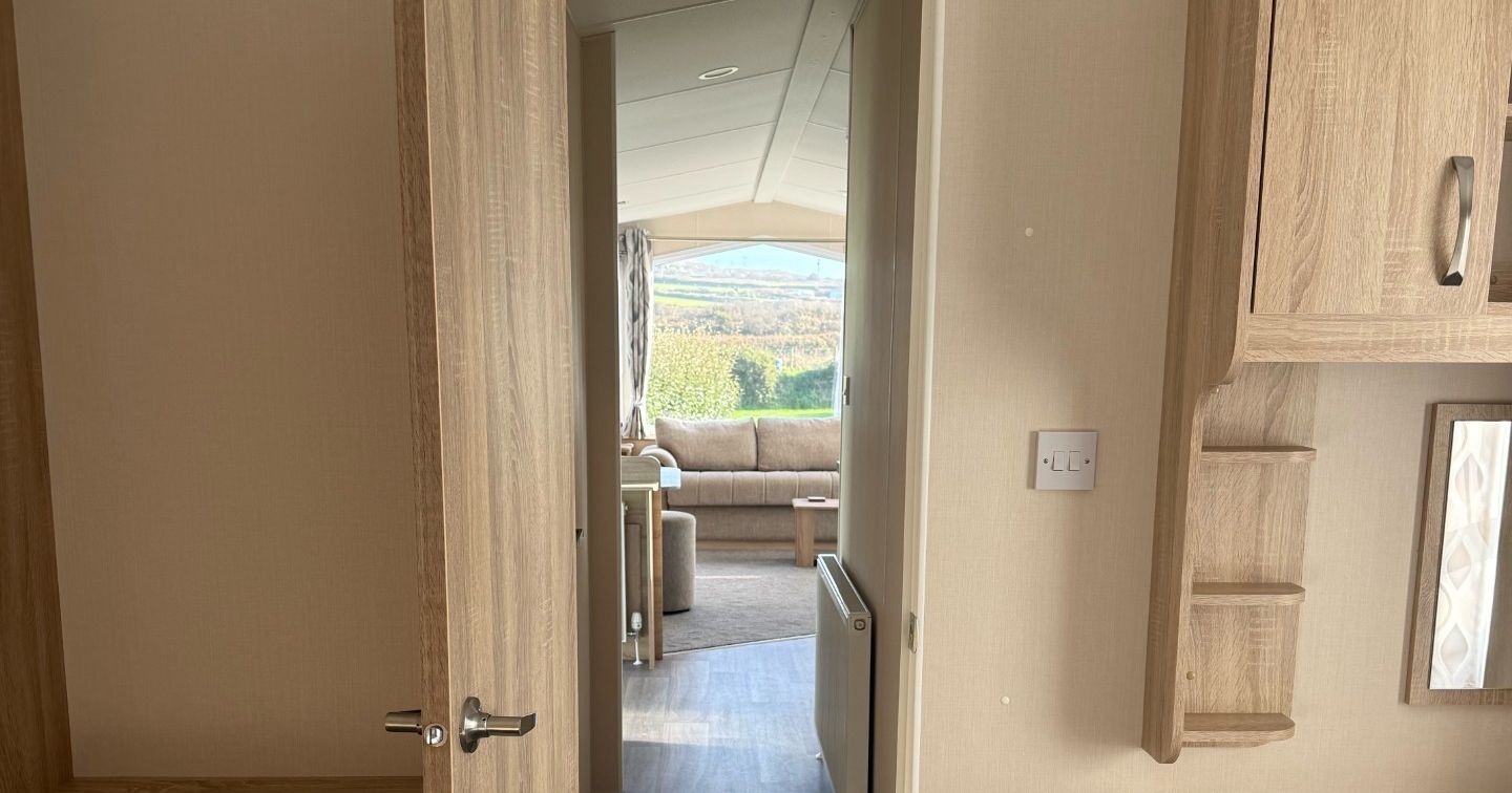 Interior view of a cozy mobile home or caravan bedroom looking through an open door into a living area with wooden furniture and a view outside.