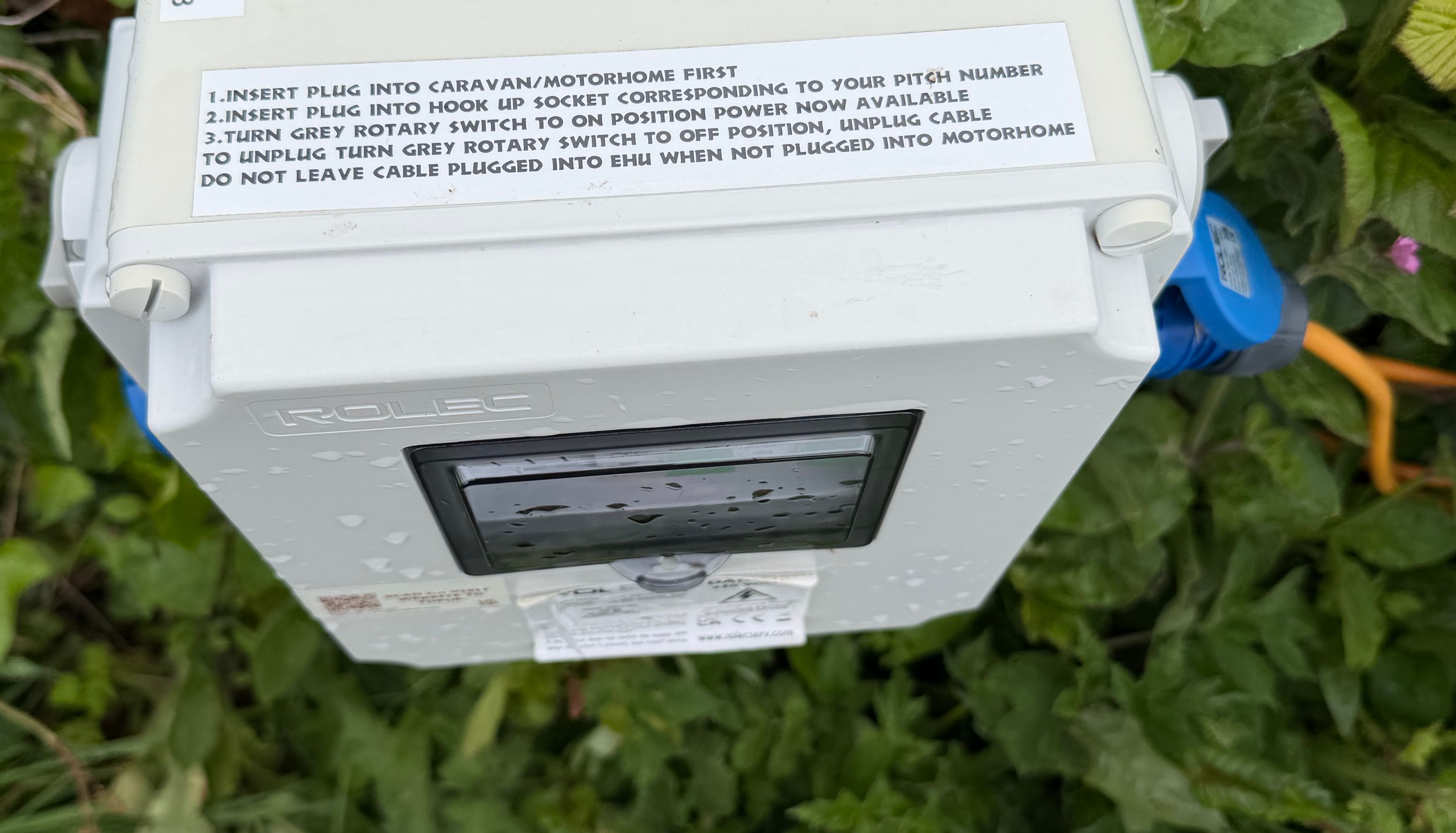 Outdoor caravan electric hook-up box with top-up instructions among green leaves