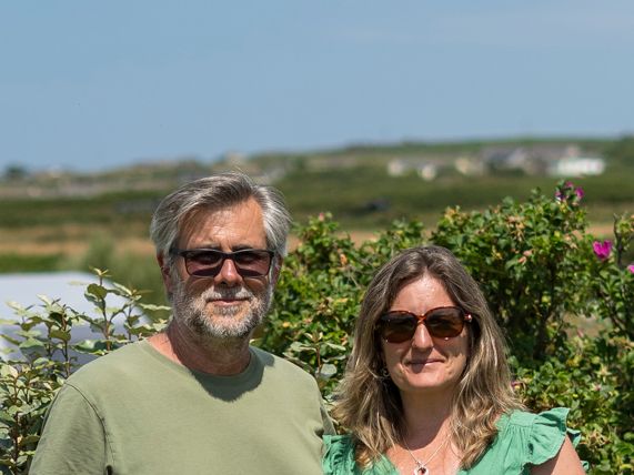 Two adults standing in front of a hedge with pink flowers at Trevaylor Caravan and Camping Park