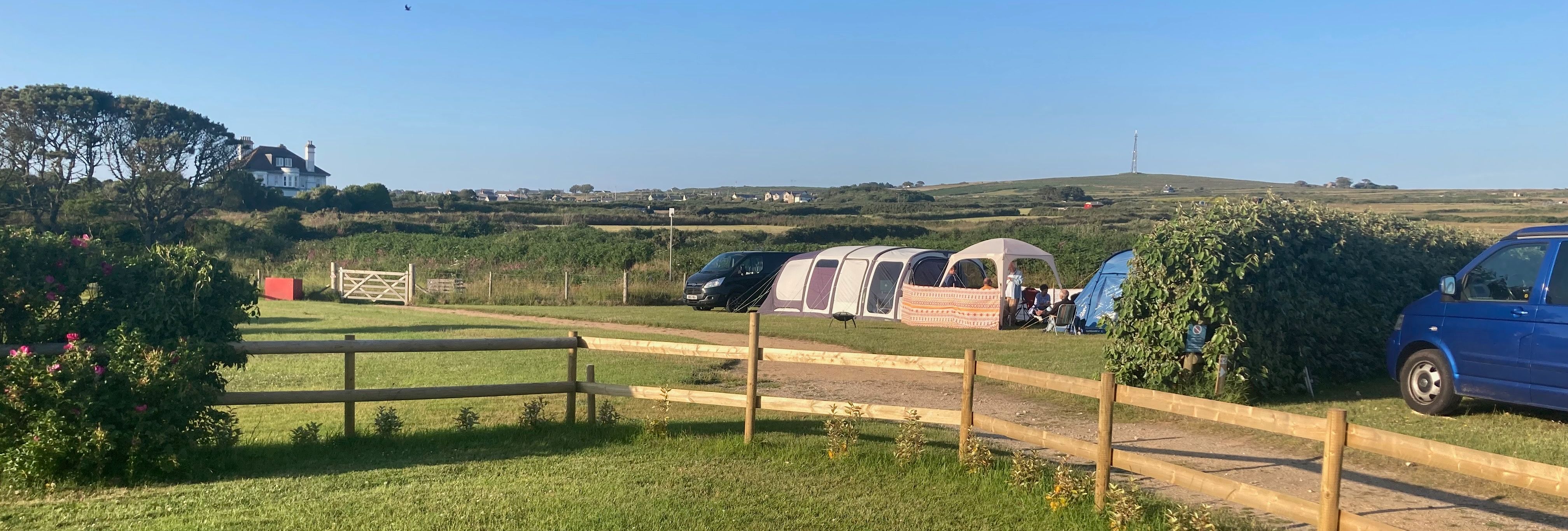 A campsite with tents and vans on a sunny day, surrounded by green fields and a wooden fence.