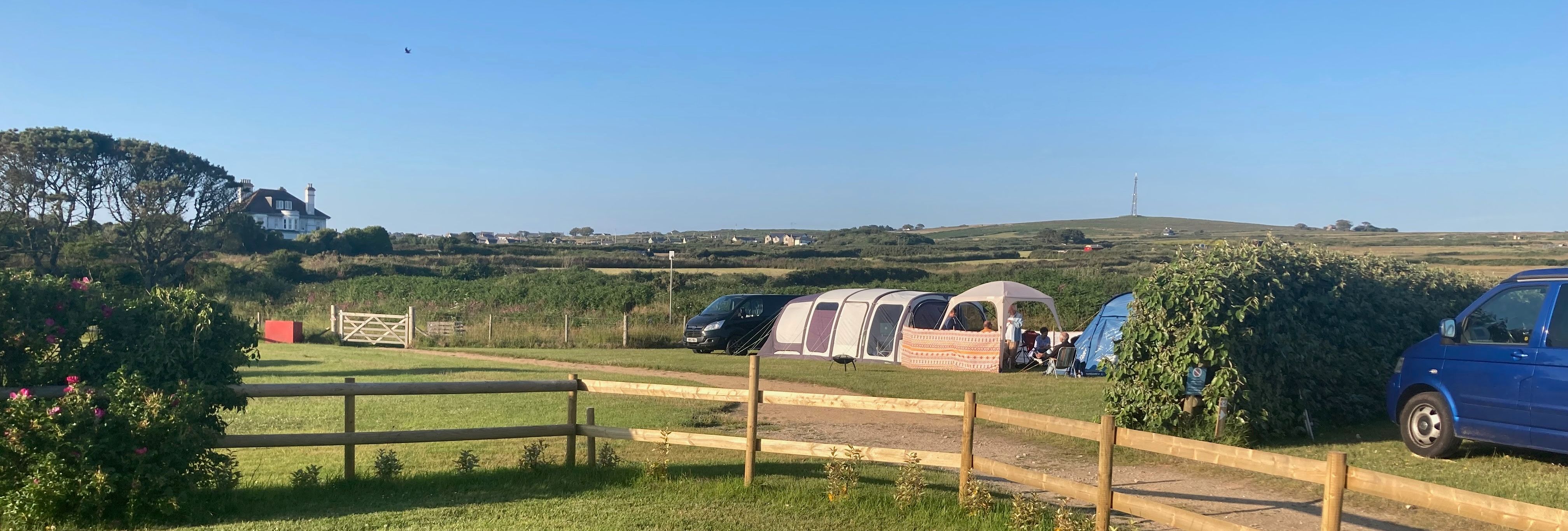 A campsite with tents and vans on a sunny day, surrounded by green fields and a wooden fence.