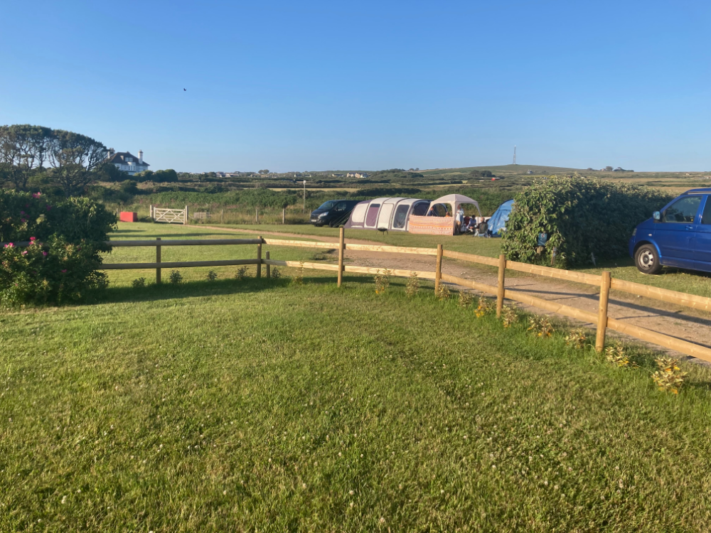 A campsite with tents and vans on a sunny day, surrounded by green fields and a wooden fence.