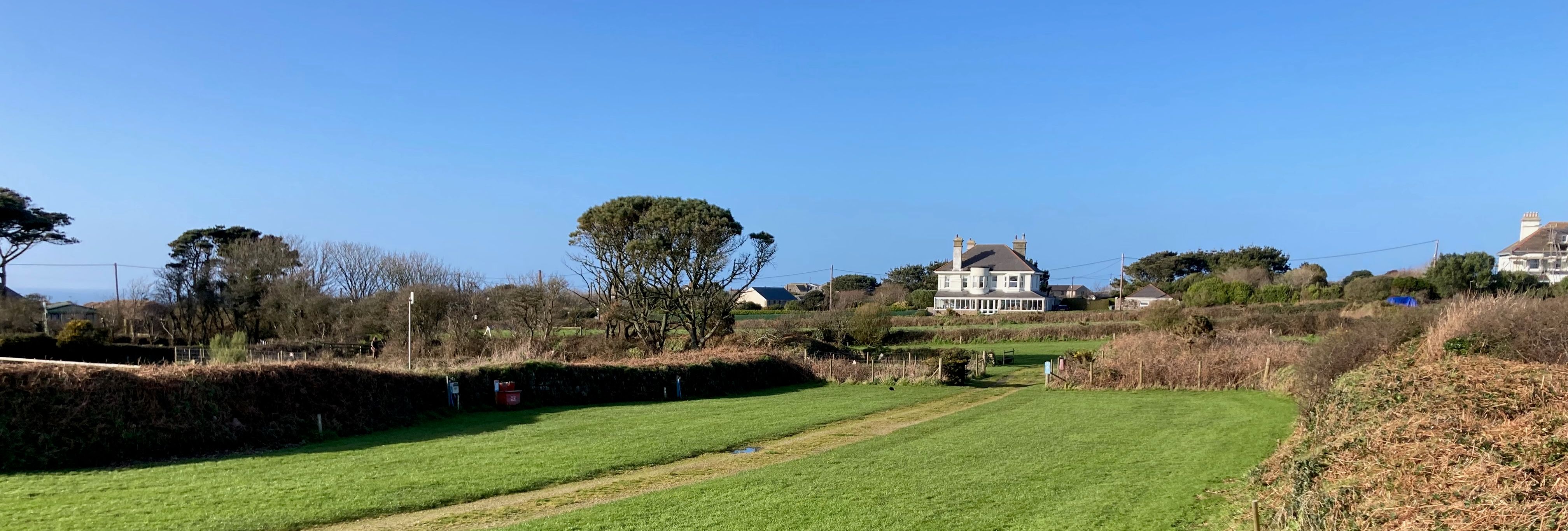 A green grassy field with a dirt path, bordered by hedges and trees, with a large house in the background under a clear blue sky.