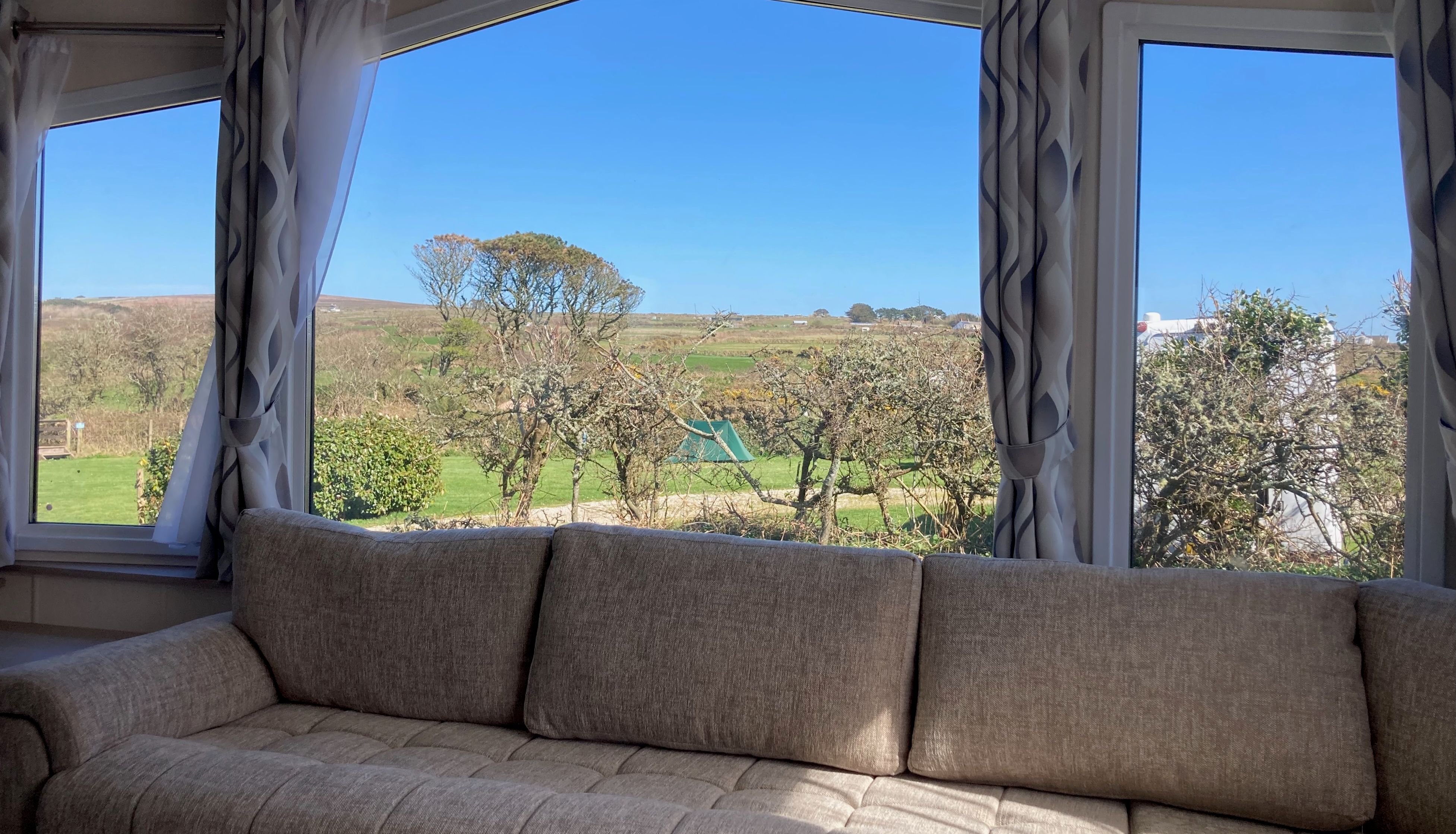 Large beige sofa positioned in front of bright windows with views of green countryside and blue sky.