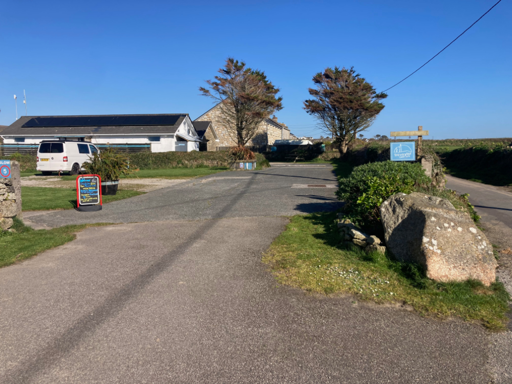 Country lane leading to a building complex, with a sign for Trevaylor Caravan & Camping Park and a van parked near a board.