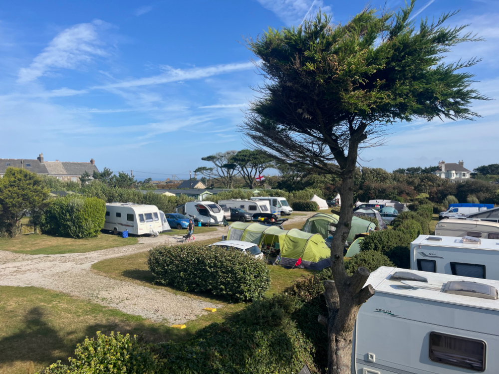 Camping site with caravans, tents and cars on hardstanding pitches surrounded by hedges and trees under a blue sky.