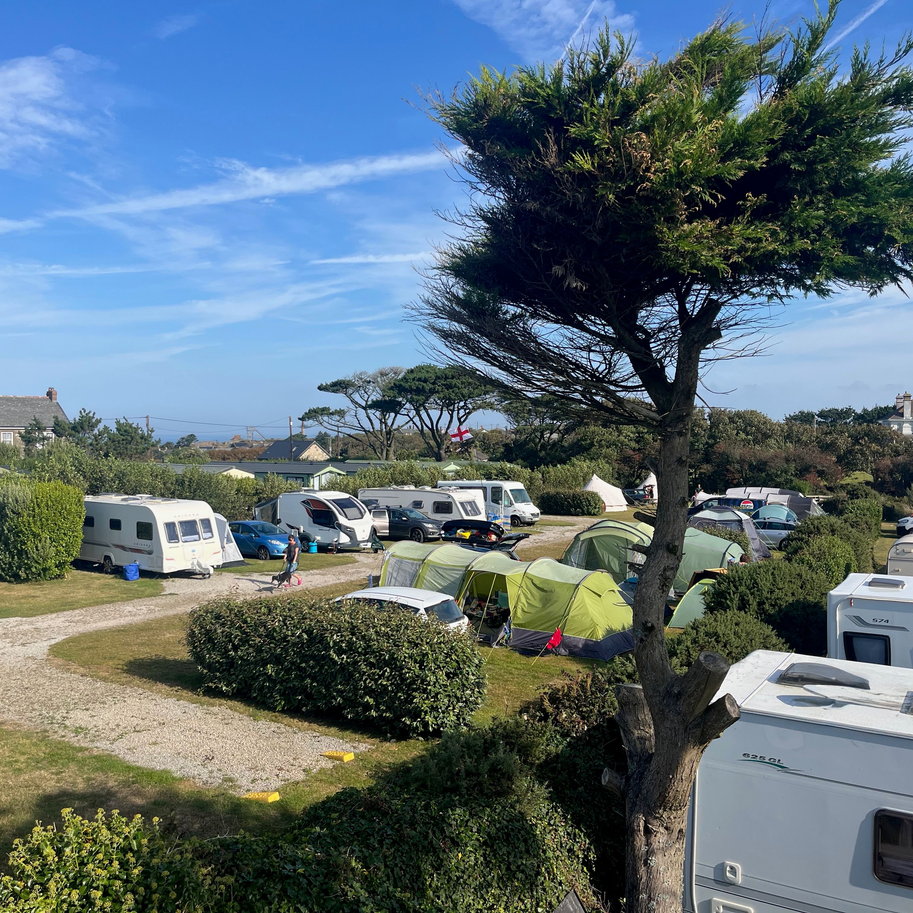 Camping site with caravans, tents and cars on hardstanding pitches surrounded by hedges and trees under a blue sky.