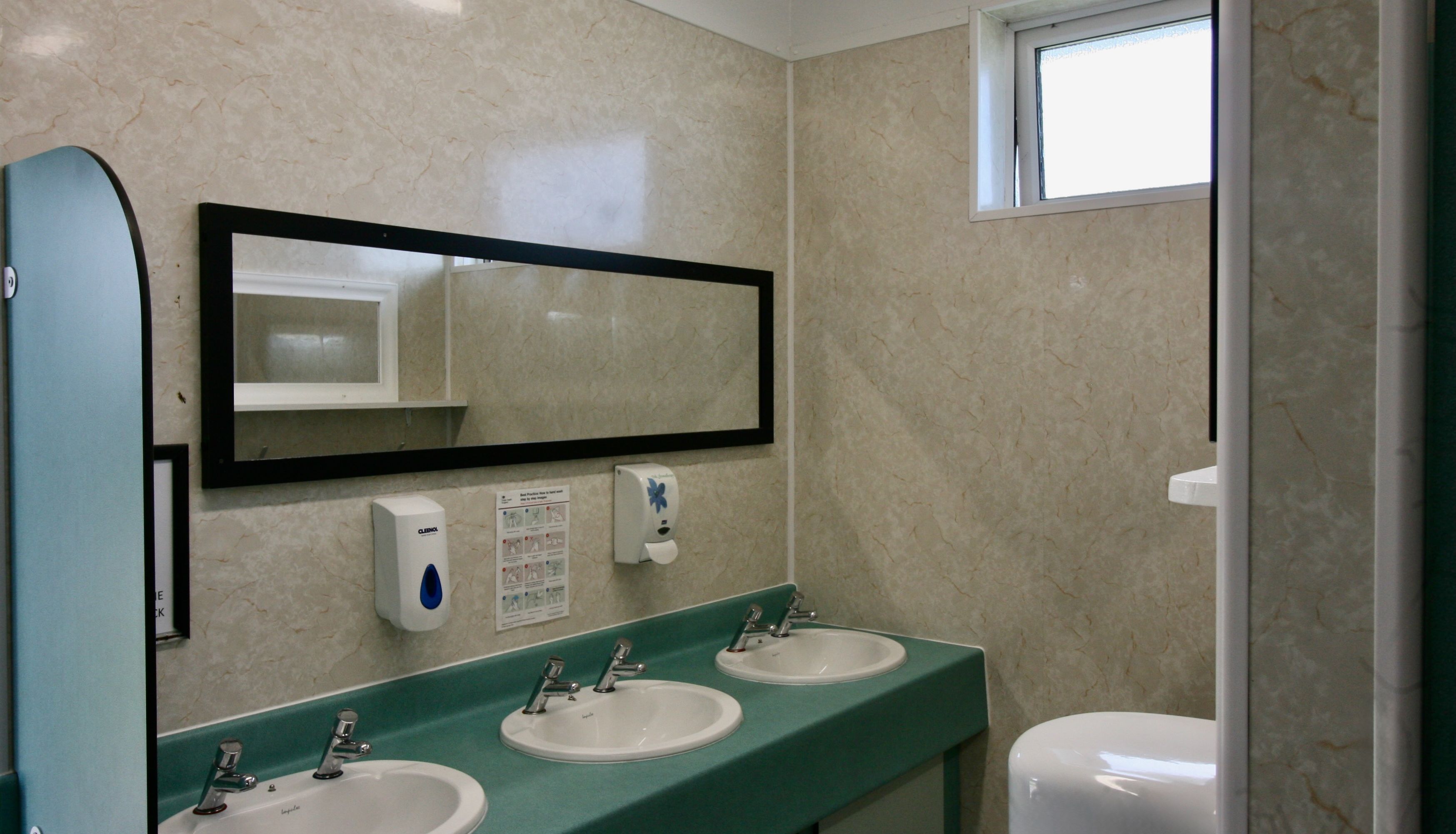 Public restroom with three sinks, soap dispensers, a rectangular mirror, and a small window.