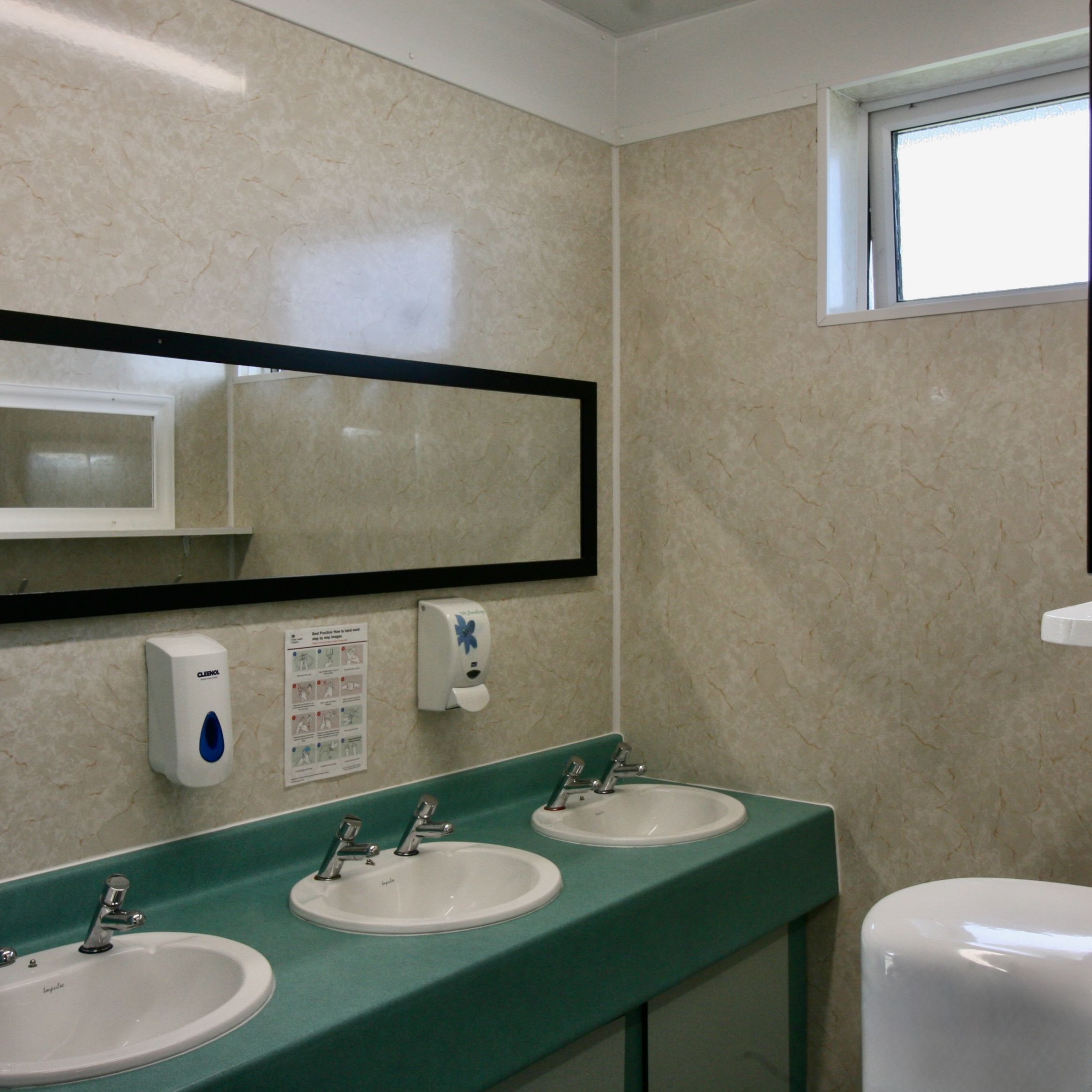 Public restroom with three sinks, soap dispensers, a rectangular mirror, and a small window.