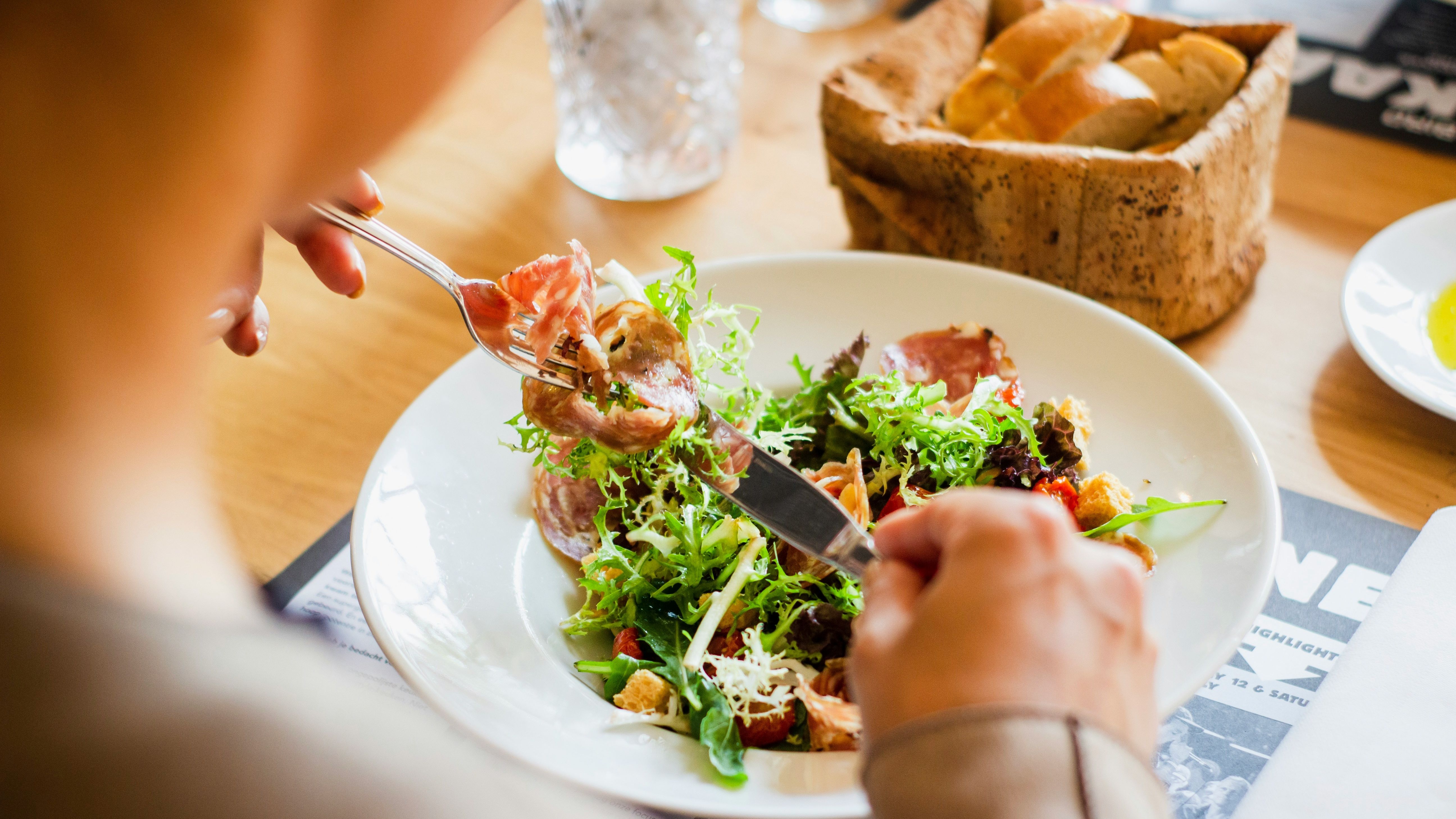 Person eating a fresh salad with cured meats using a fork and knife at a wooden table.