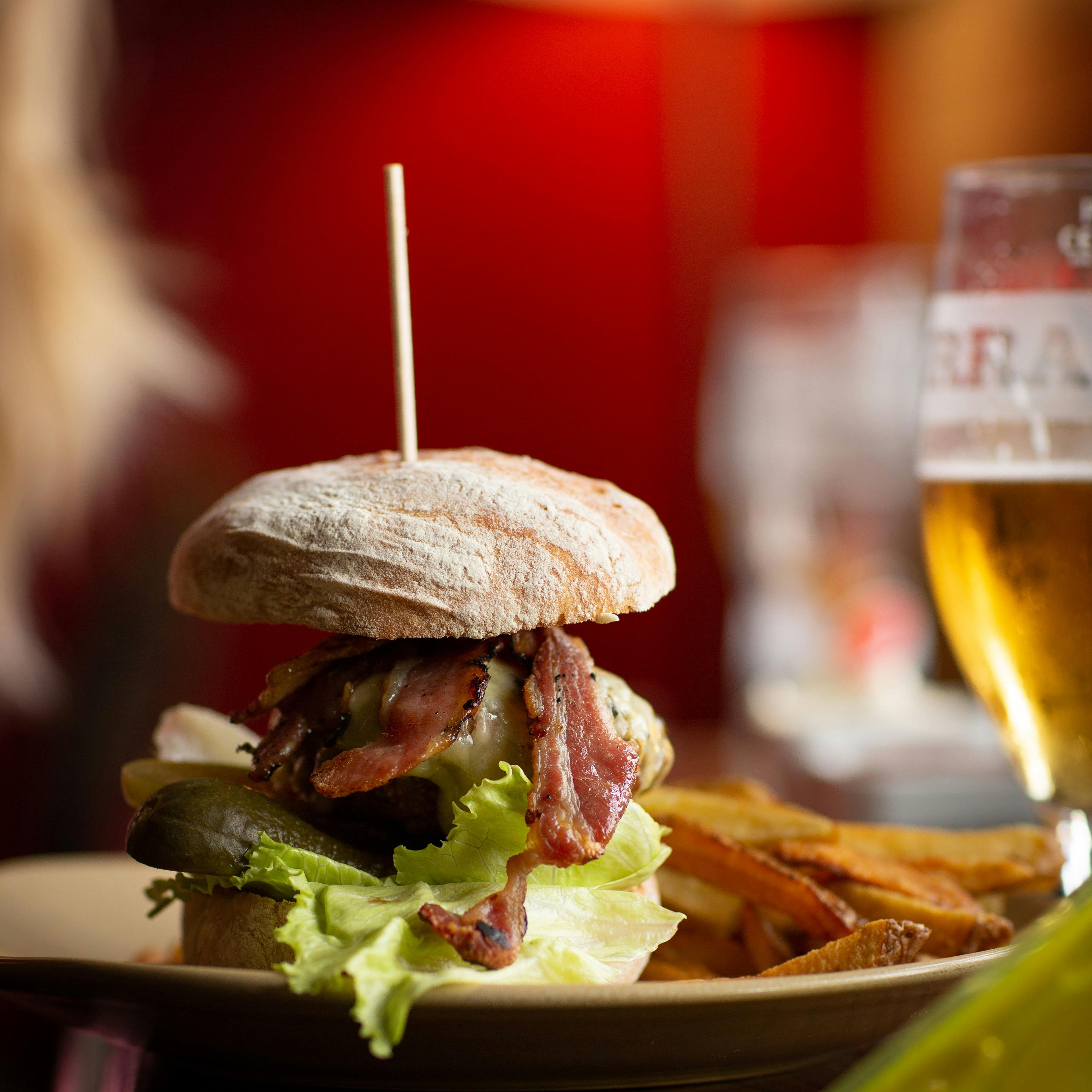 Close-up of a gourmet burger with bacon, lettuce, pickles, and a rustic bun served with fries and a glass of beer.