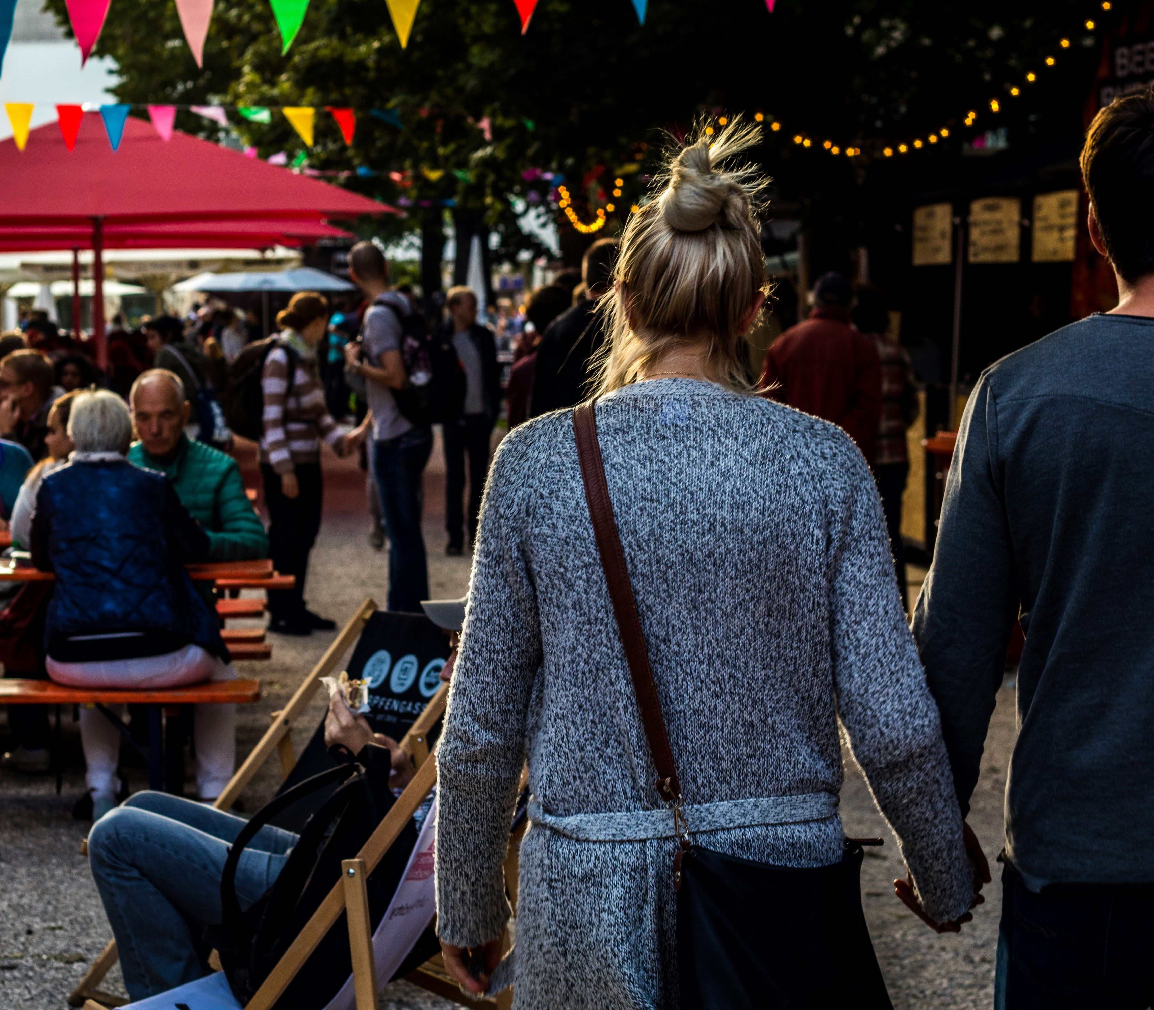 Couple holding hands walking through a lively outdoor festival with people sitting at tables and colorful bunting overhead.