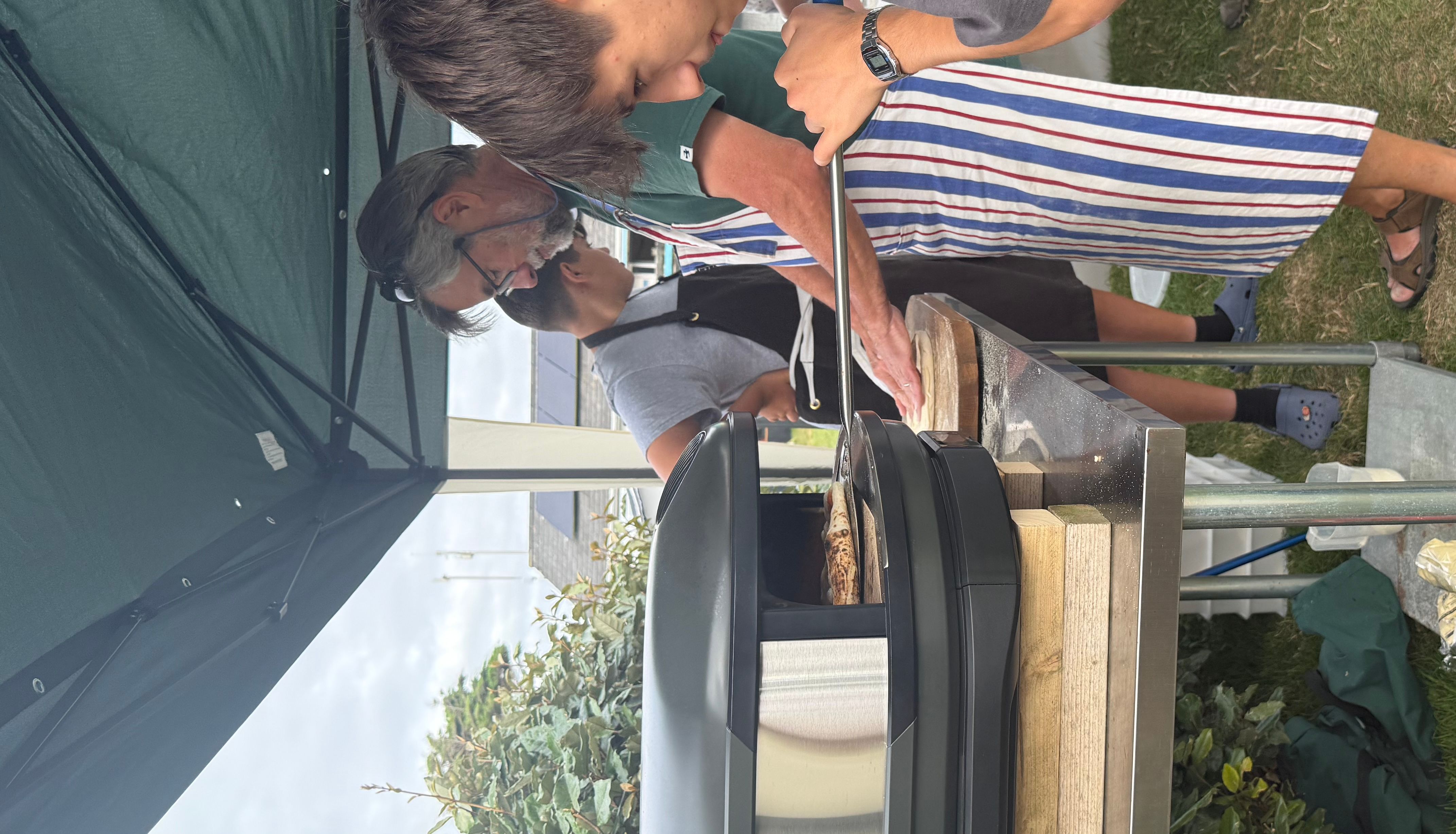 Group of people making pizza outdoors under a canopy tent with a portable pizza oven.
