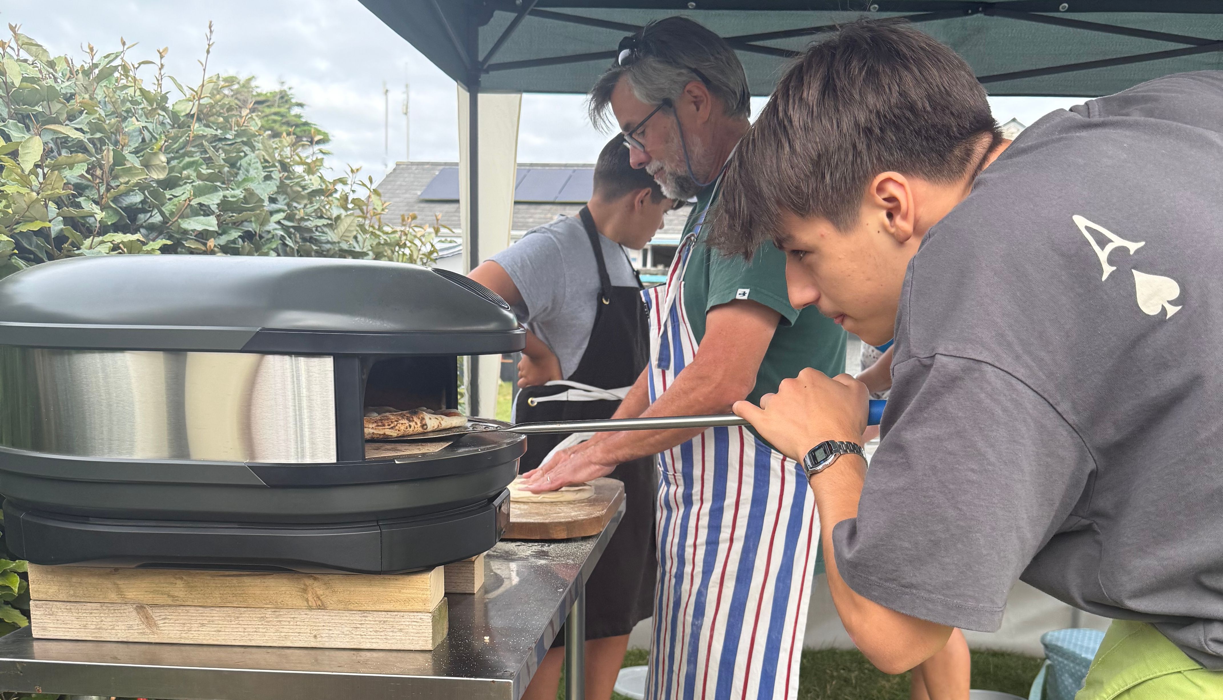 Group of people making pizza outdoors under a canopy tent with a portable pizza oven.