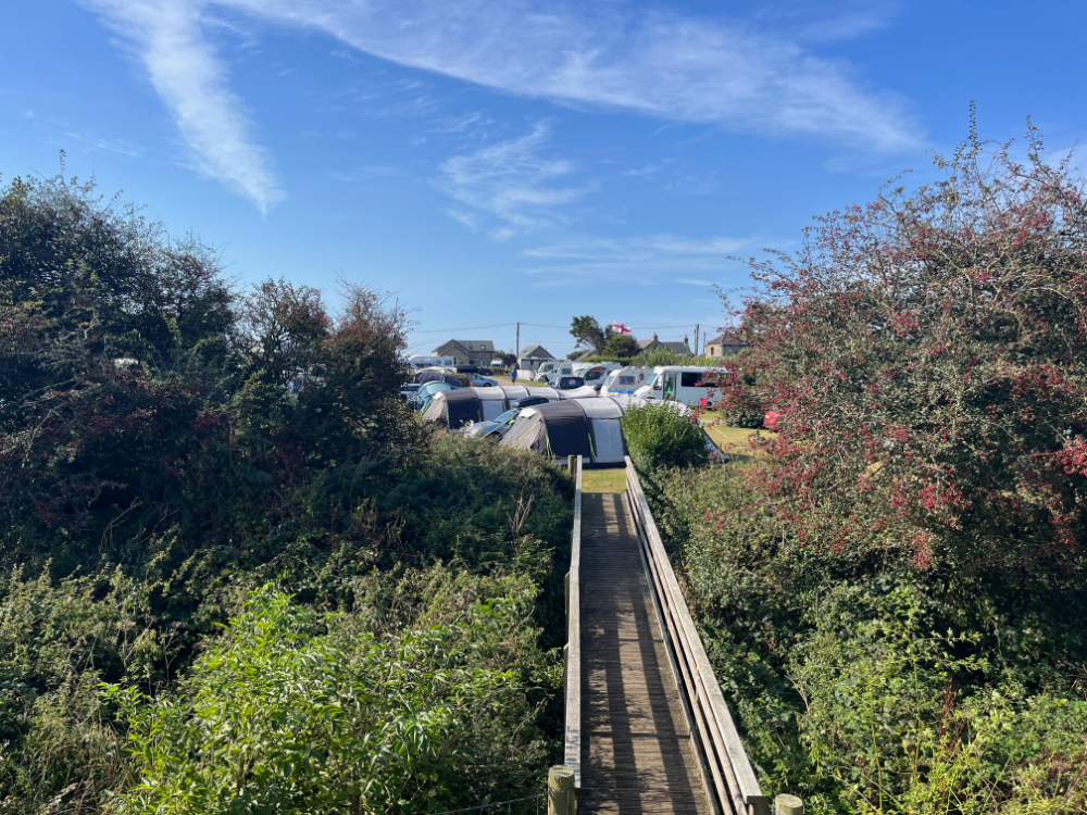 A wooden footbridge leading through bushes to a camping area with tents and campervans under a bright blue sky.