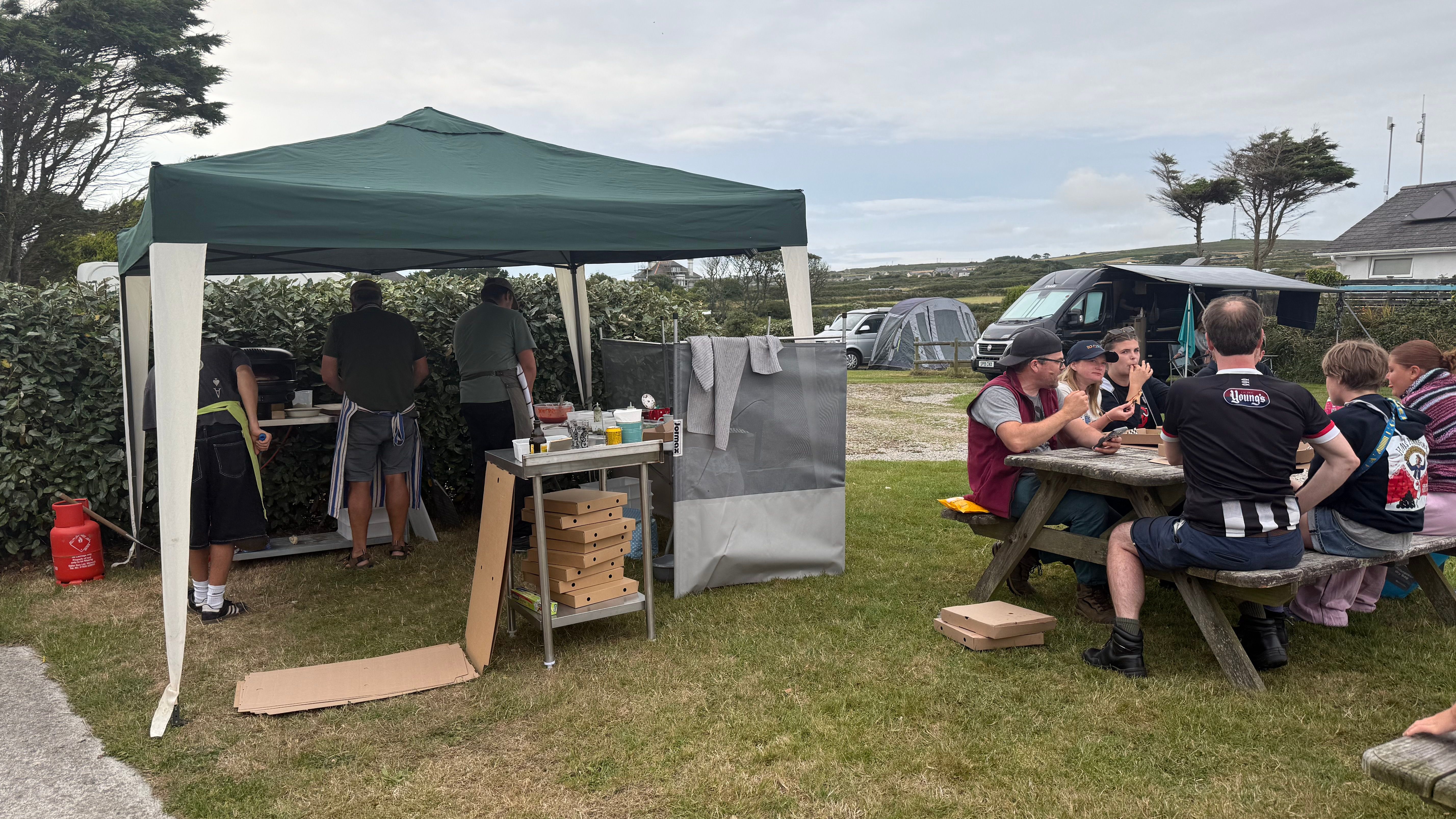 A group of people eating at a picnic table near a tent where others are preparing food outdoors.