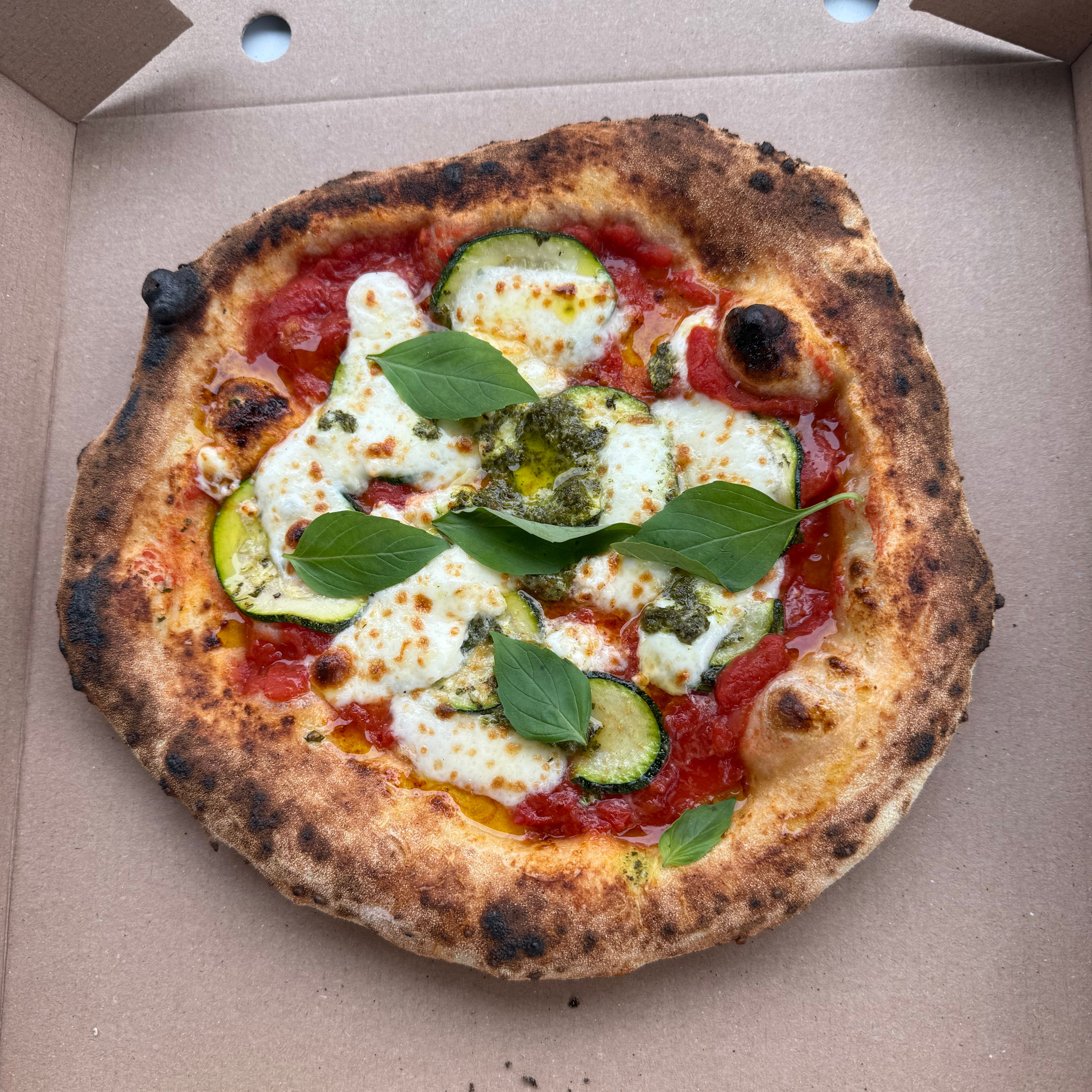 A freshly baked pizza in a cardboard box topped with melted cheese, zucchini slices, tomato sauce, and fresh basil leaves.
