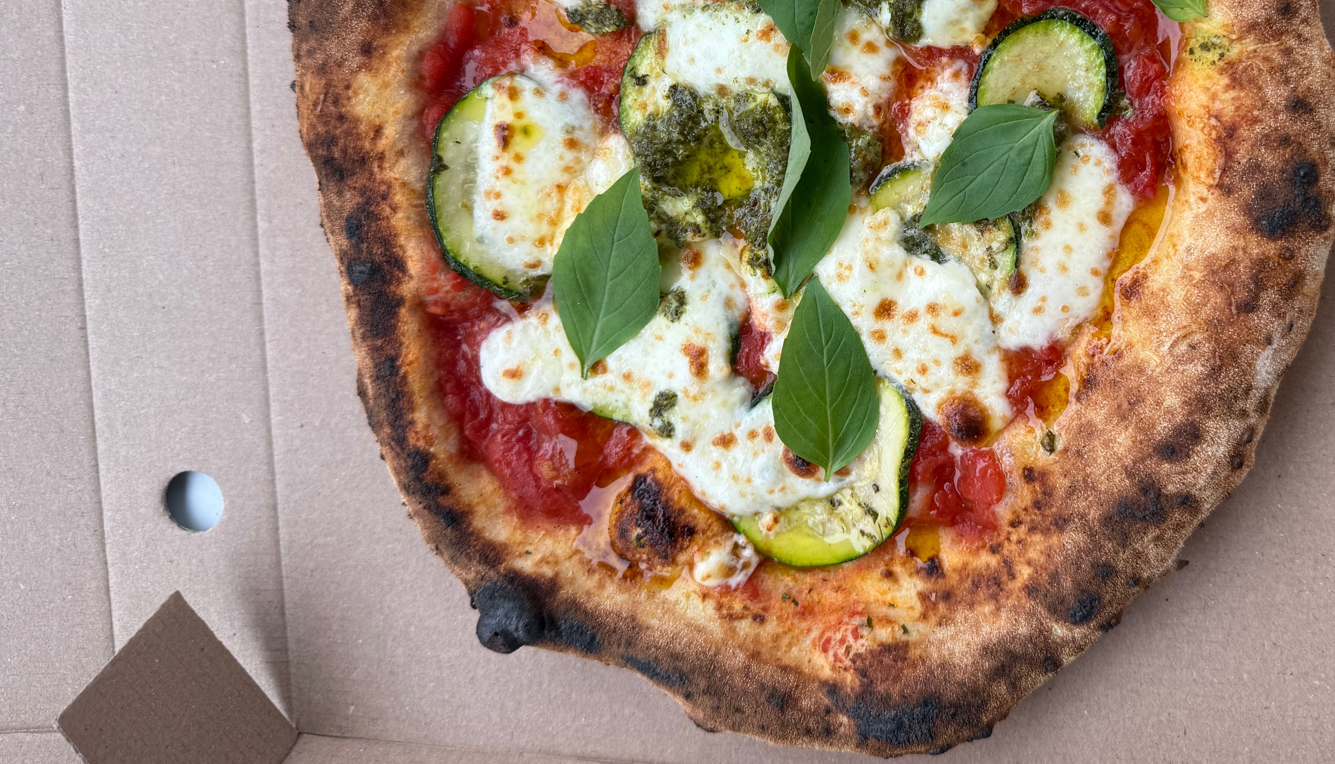 A freshly baked pizza in a cardboard box topped with melted cheese, zucchini slices, tomato sauce, and fresh basil leaves.