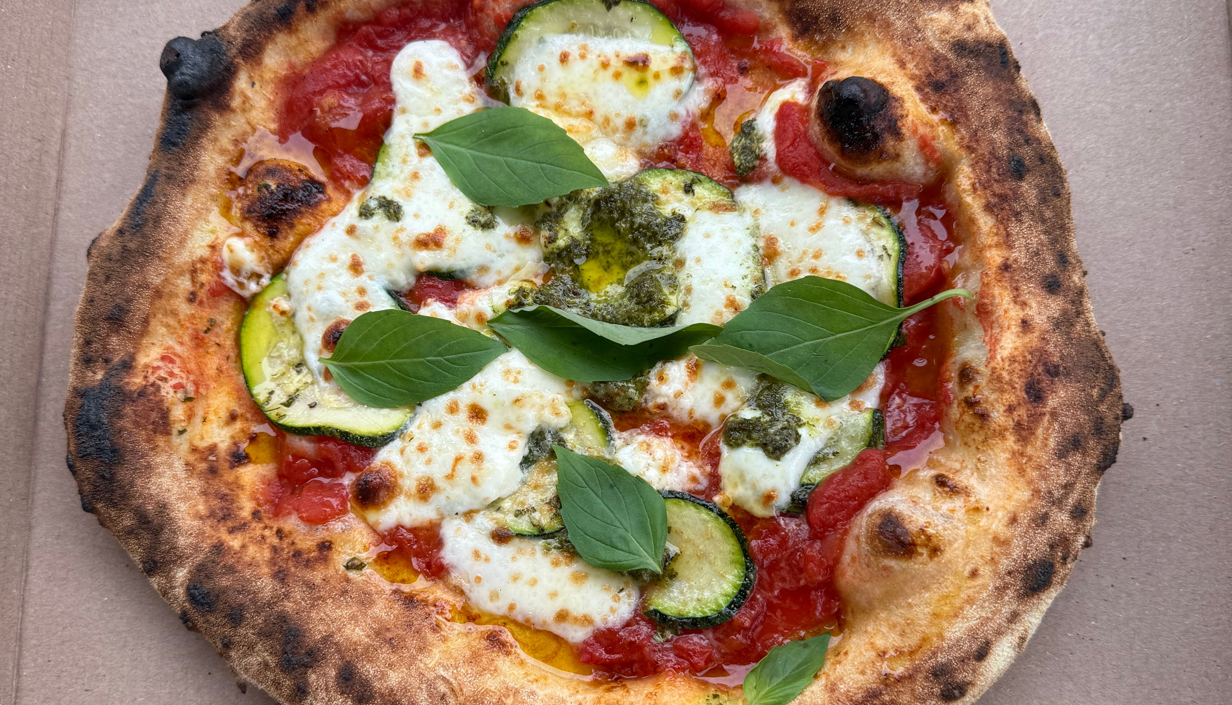 A freshly baked pizza in a cardboard box topped with melted cheese, zucchini slices, tomato sauce, and fresh basil leaves.
