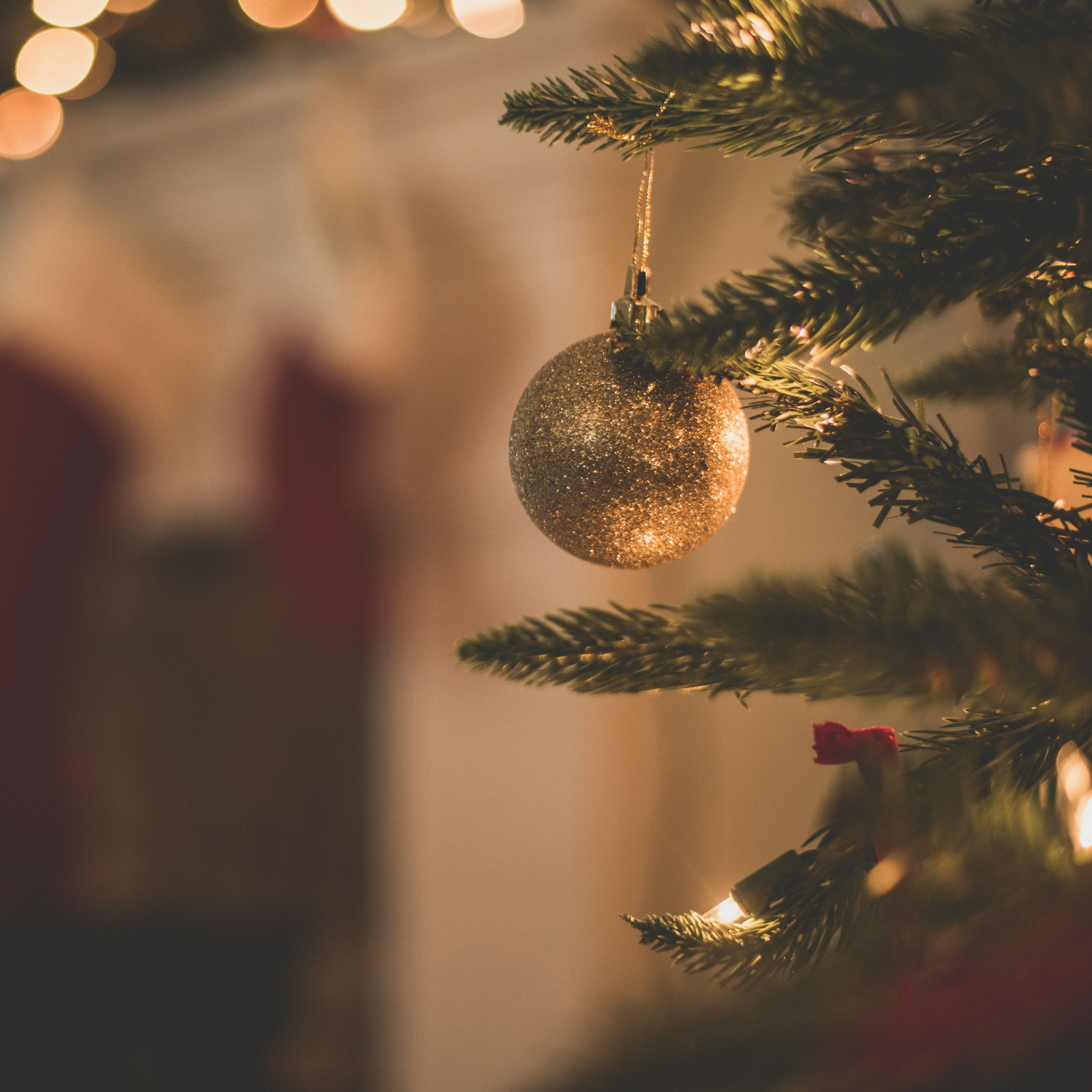 Close-up of a glittery gold Christmas ornament hanging on a decorated Christmas tree with blurred lights and stockings in the background.