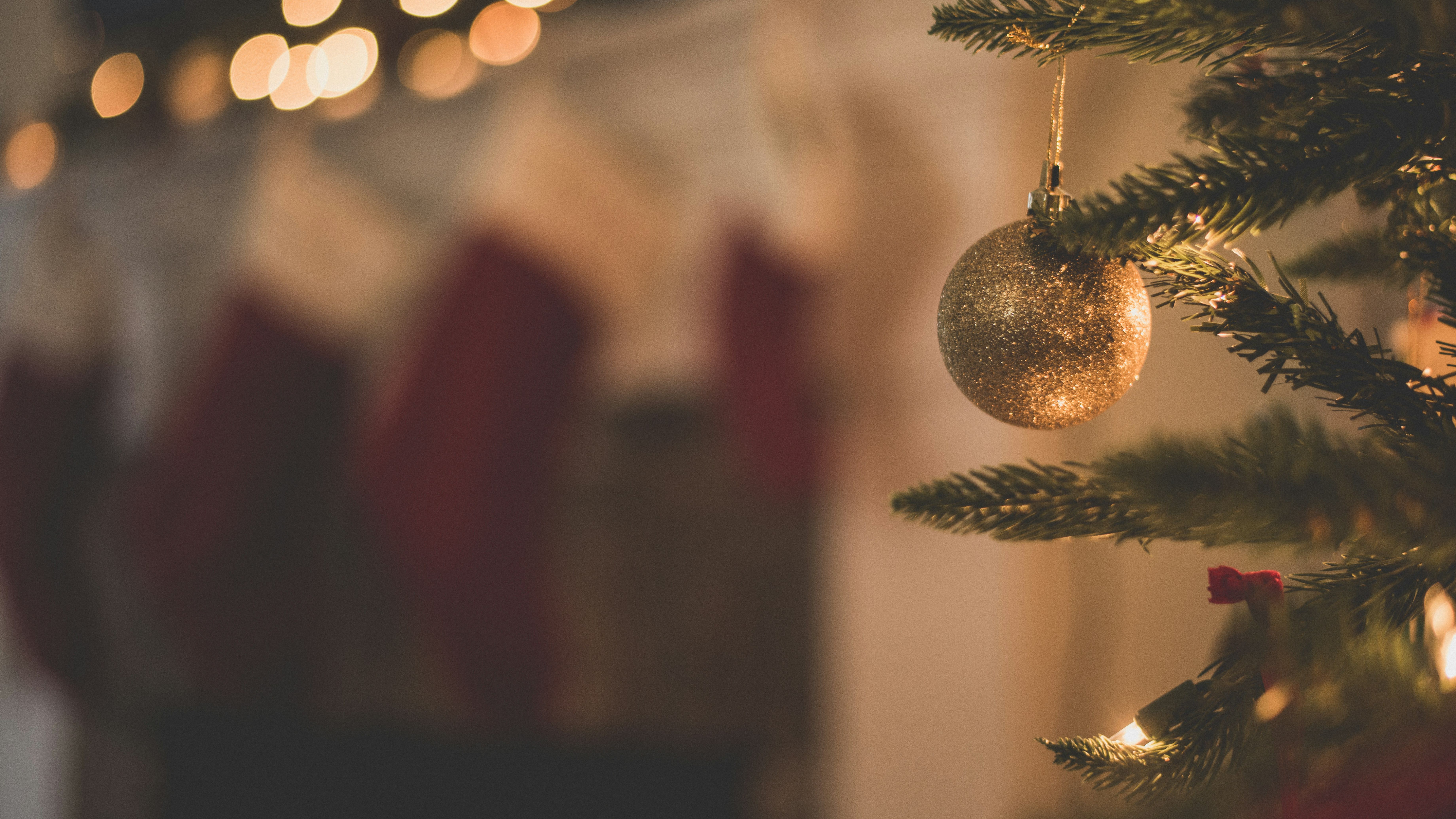 Close-up of a glittery gold Christmas ornament hanging on a decorated Christmas tree with blurred lights and stockings in the background.