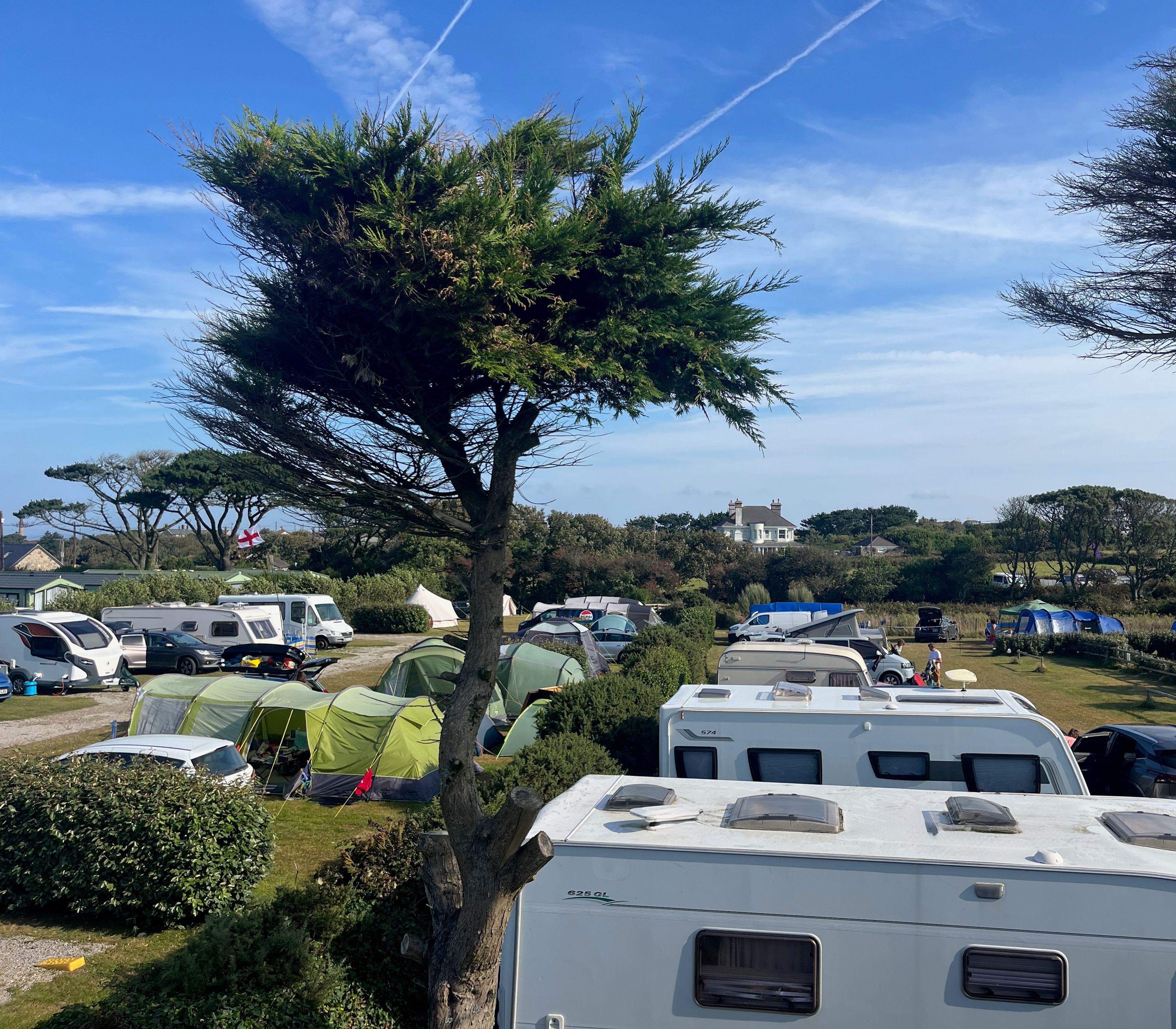 A campsite with caravans, tents, and vehicles under blue skies with scattered clouds and trees.