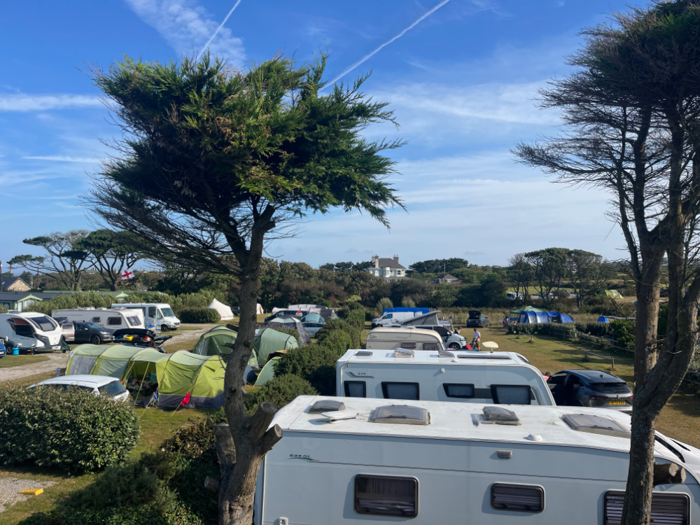A campsite with caravans, tents, and vehicles under blue skies with scattered clouds and trees.