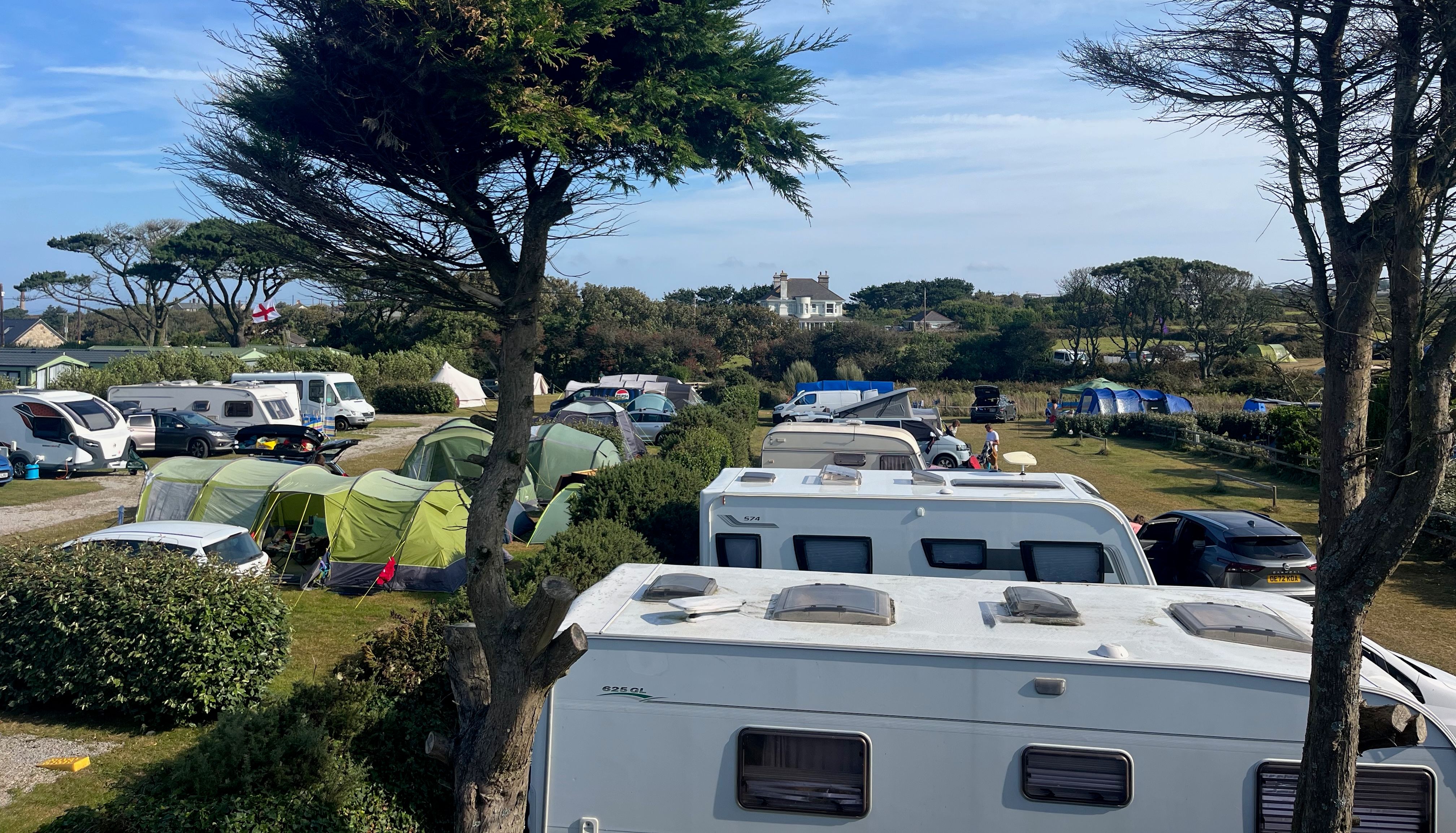 A campsite with caravans, tents, and vehicles under blue skies with scattered clouds and trees.