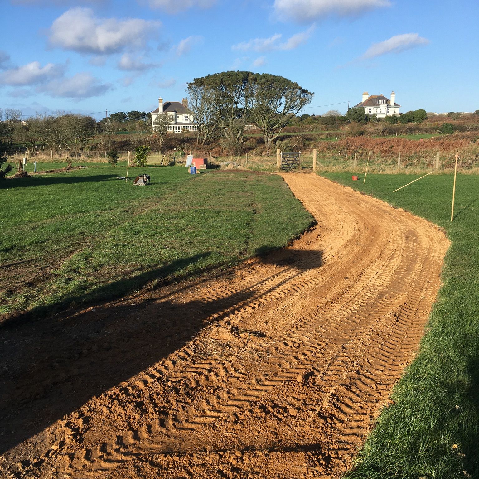 A dirt path under construction runs through a grassy field with houses and trees in the background.
