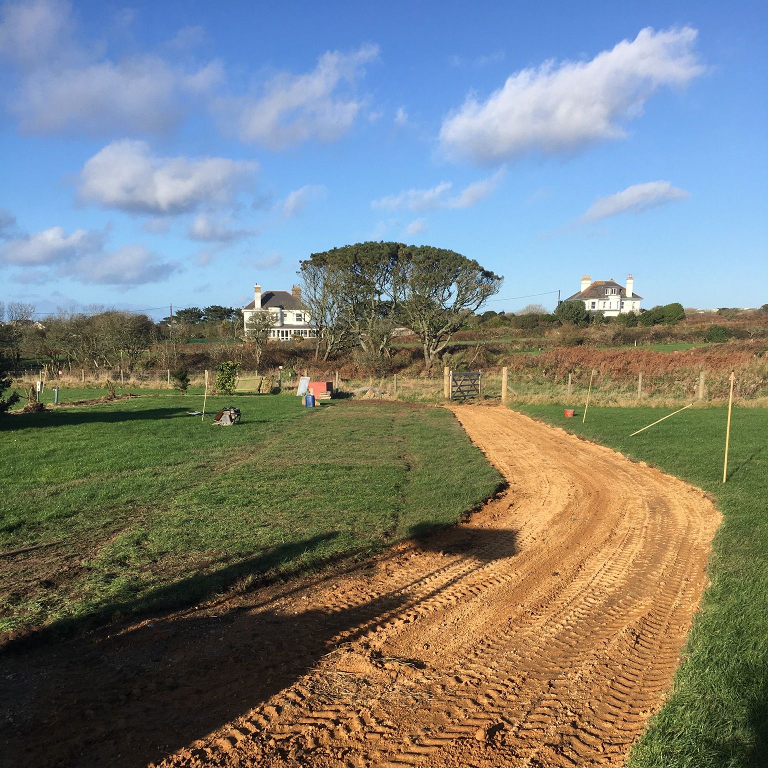 A dirt path under construction runs through a grassy field with houses and trees in the background.