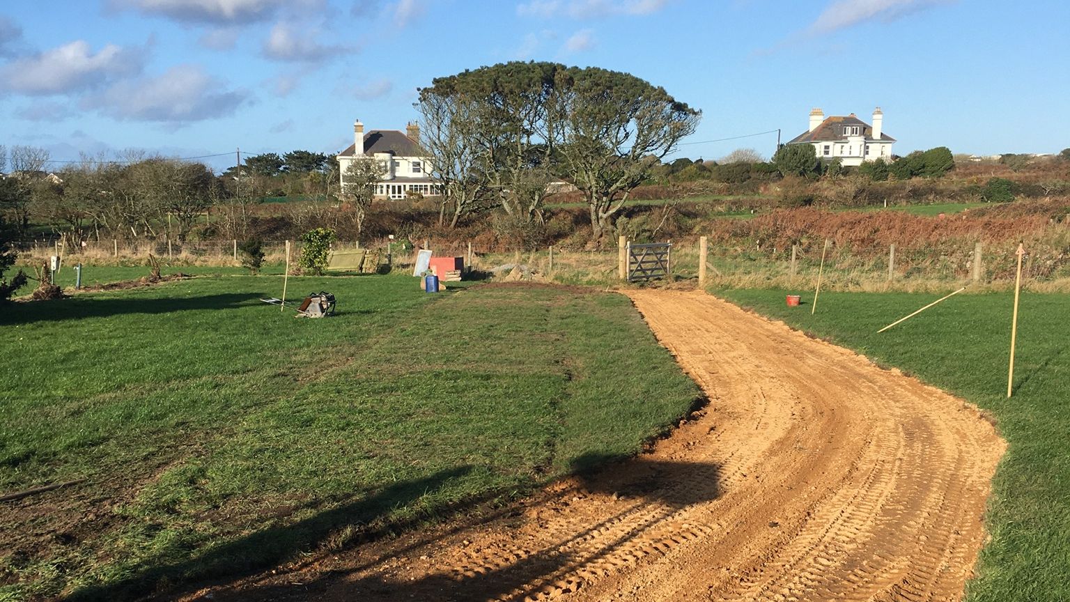A dirt path under construction runs through a grassy field with houses and trees in the background.